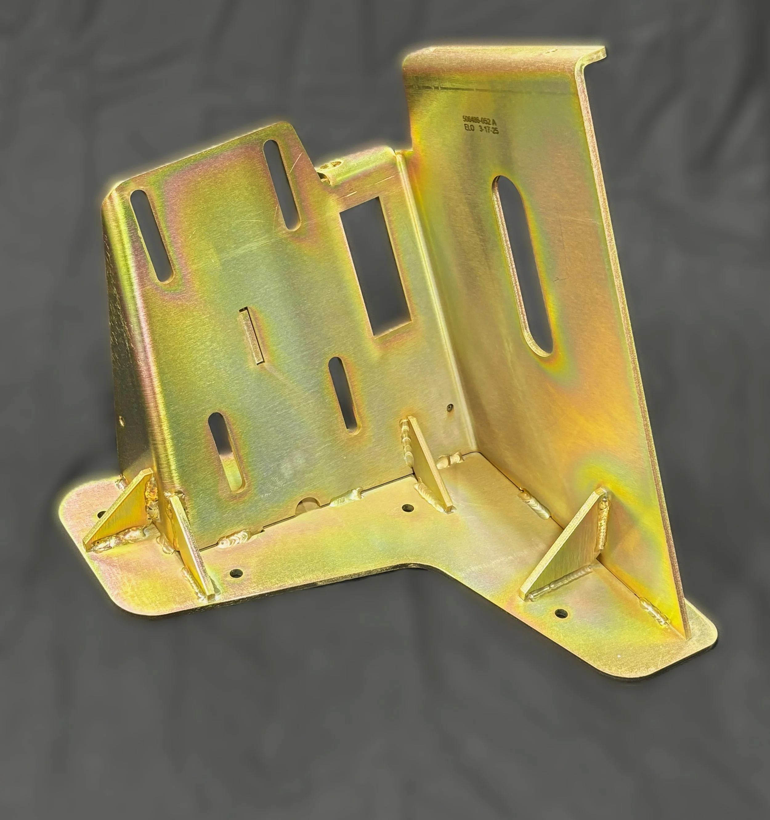 A yellowish metallic question mark-shaped bracket with multiple slots and welded joints, placed on a dark surface.