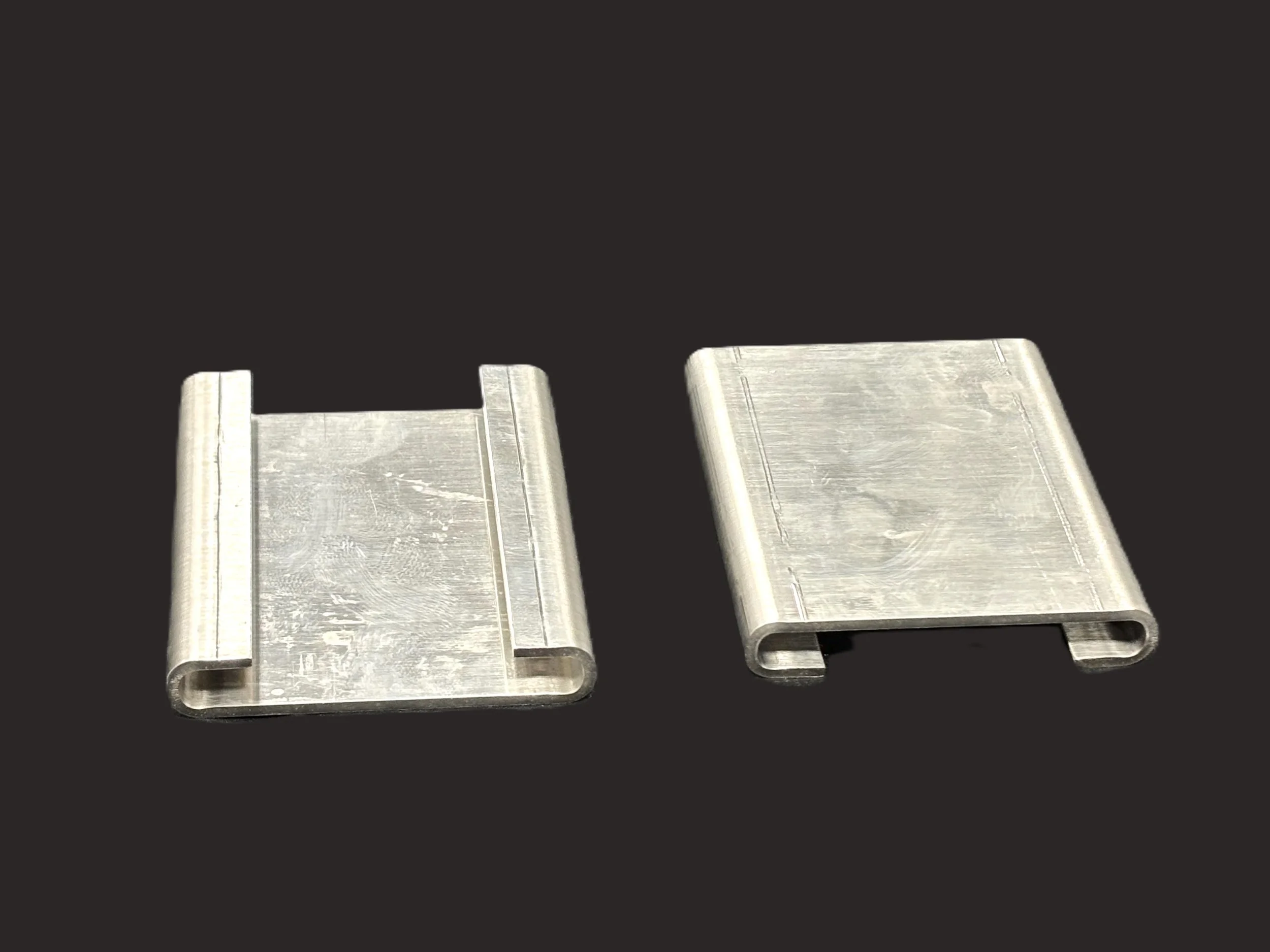 Two metallic brackets with a rectangular shape, bent at the edges, placed on a black surface.