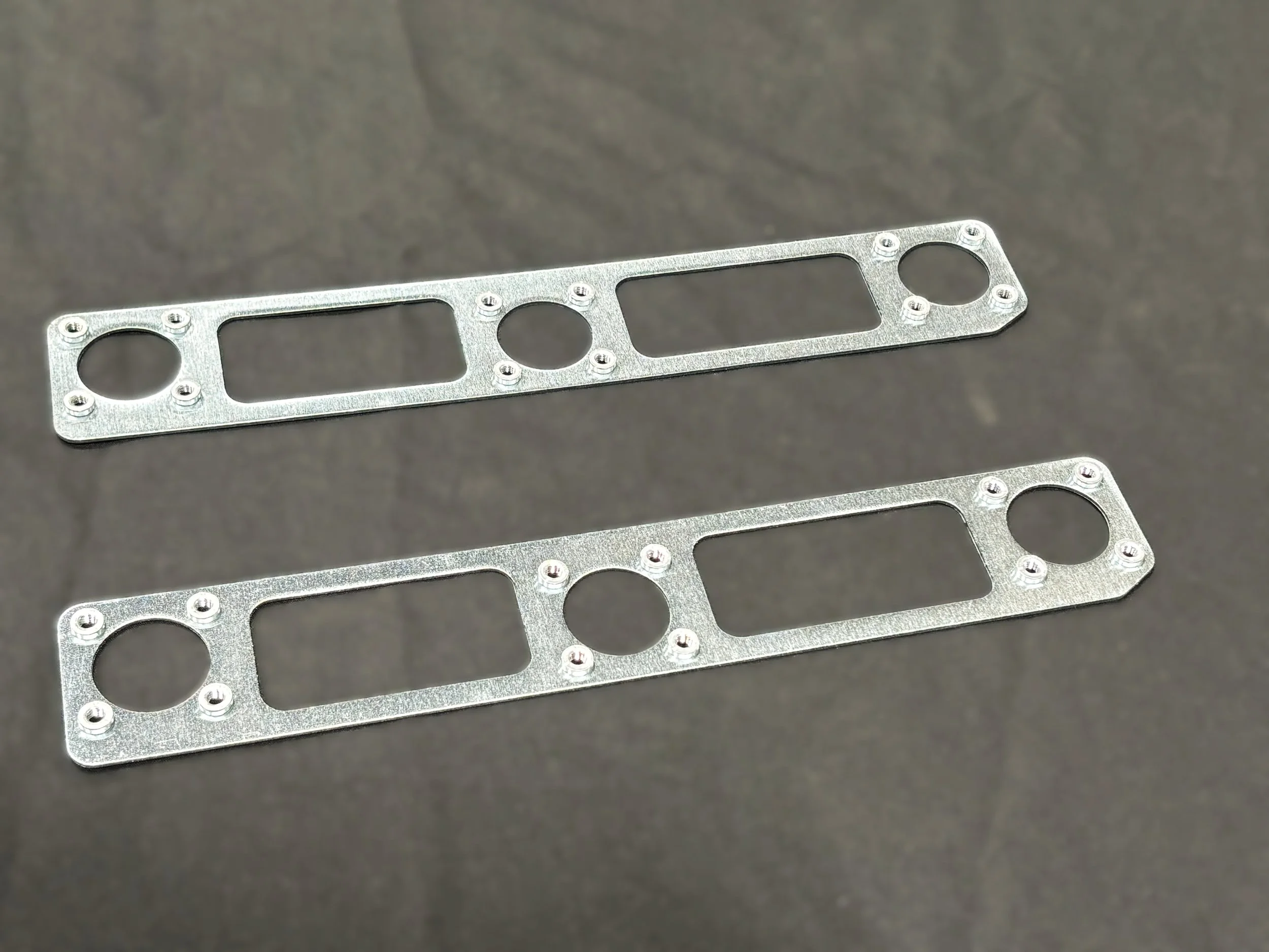 Two metallic gasket plates with circular and rectangular openings, placed on a dark surface.