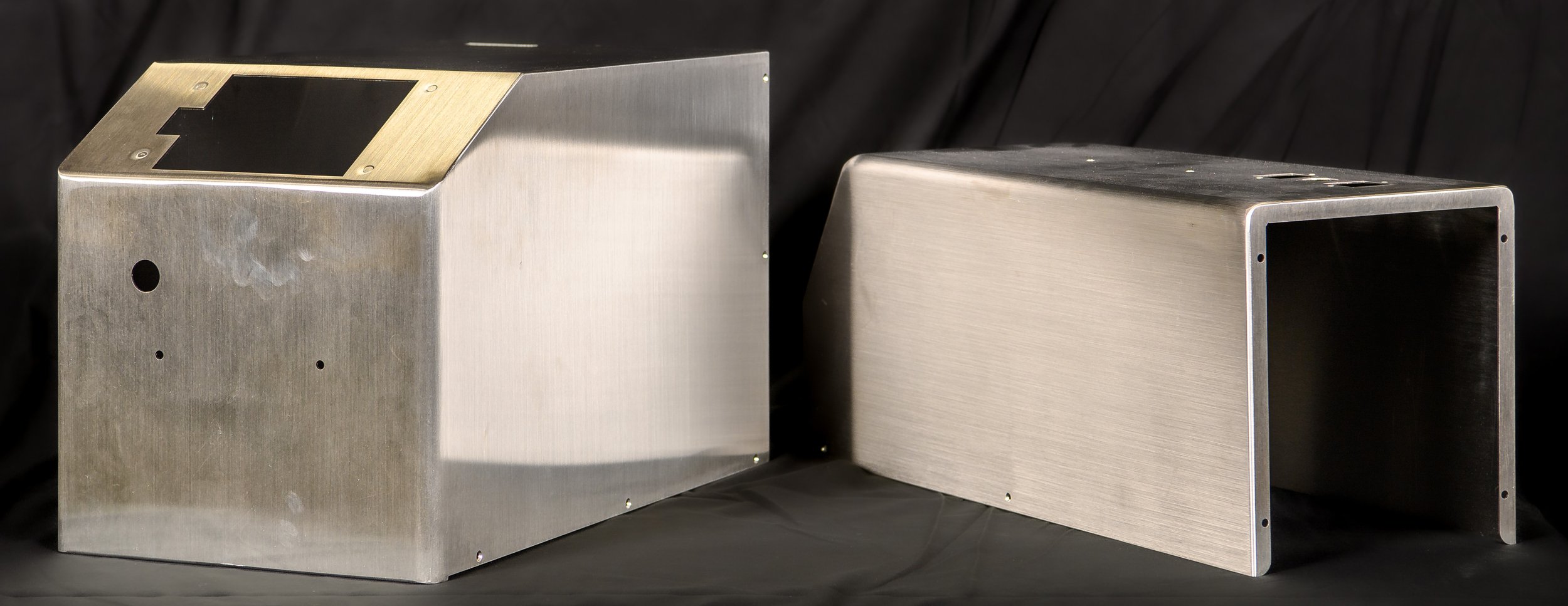 Two metallic rectangular enclosures, one with a black square panel on top and the other with an open front, placed on a dark surface against a black background.
