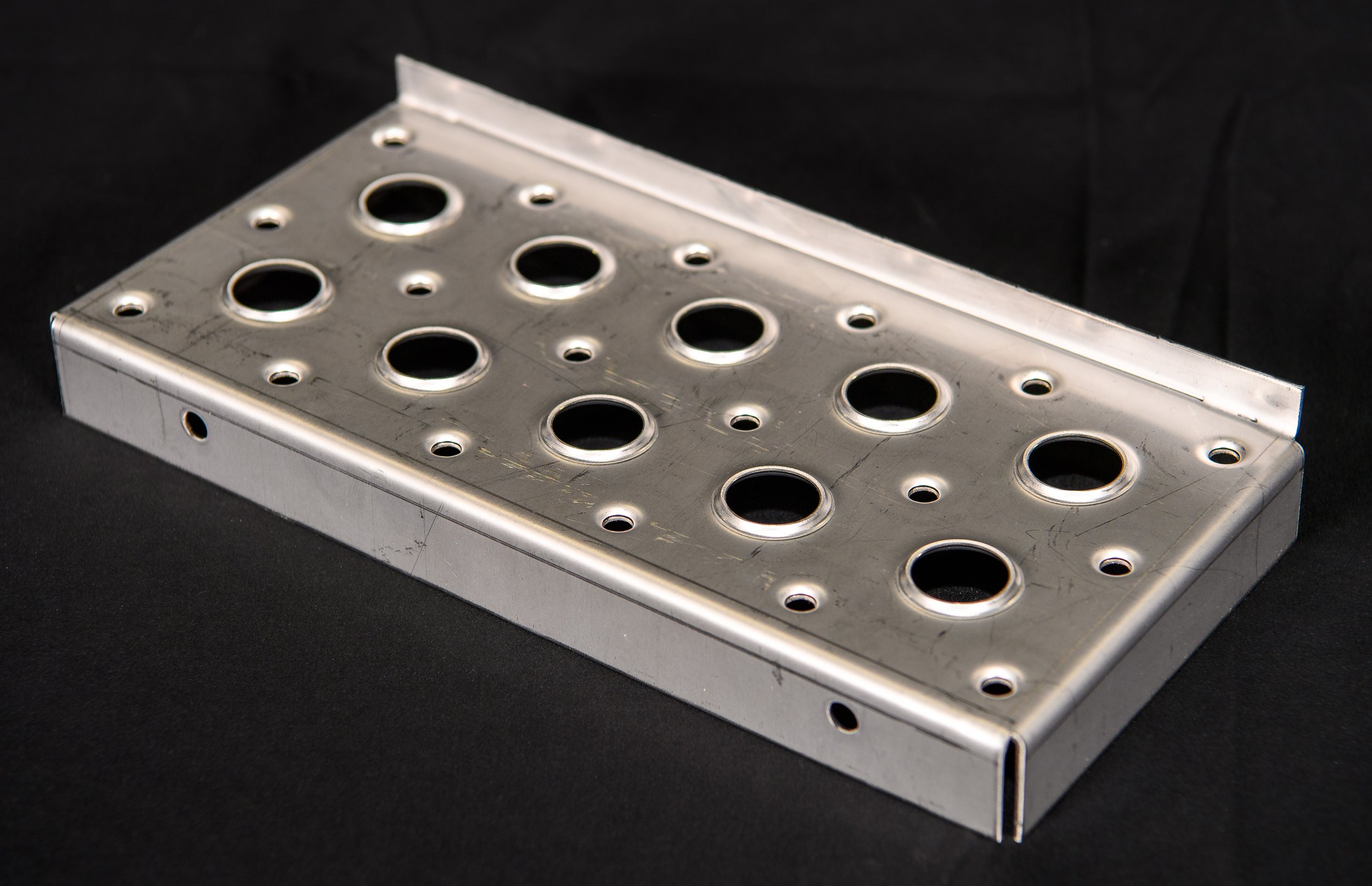 A silver metal rectangular object with multiple circular holes of various sizes, placed on a black surface.
