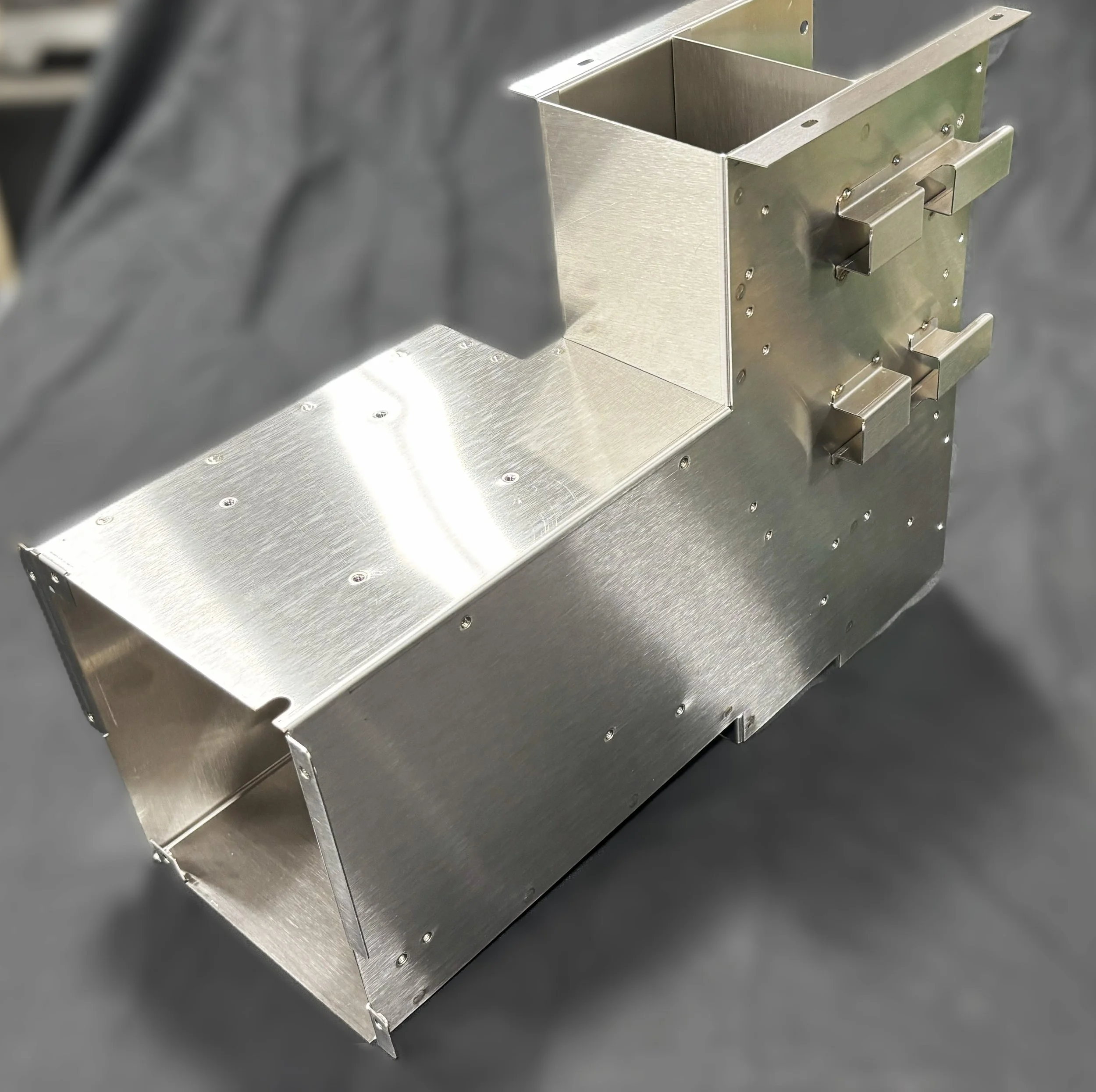 A shiny, metallic, L-shaped machinery component with multiple bolts and protruding brackets, set against a dark, blurry background.