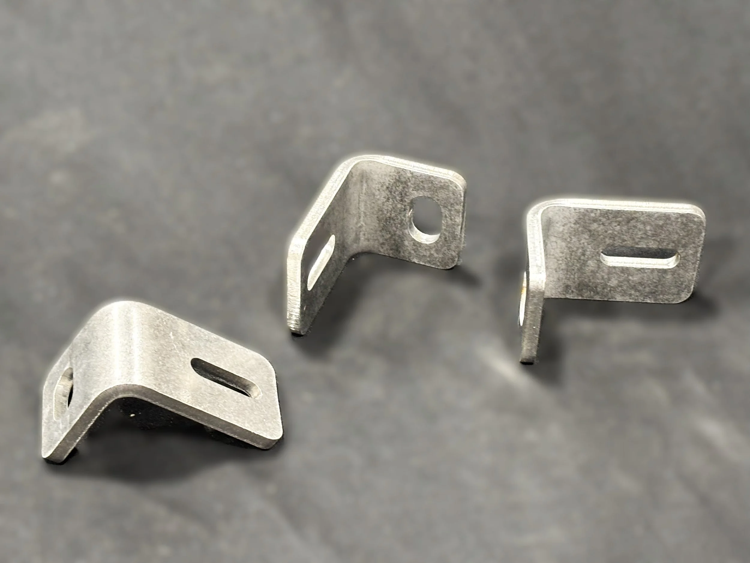 Three metal brackets with slots and holes, arranged on a gray surface.