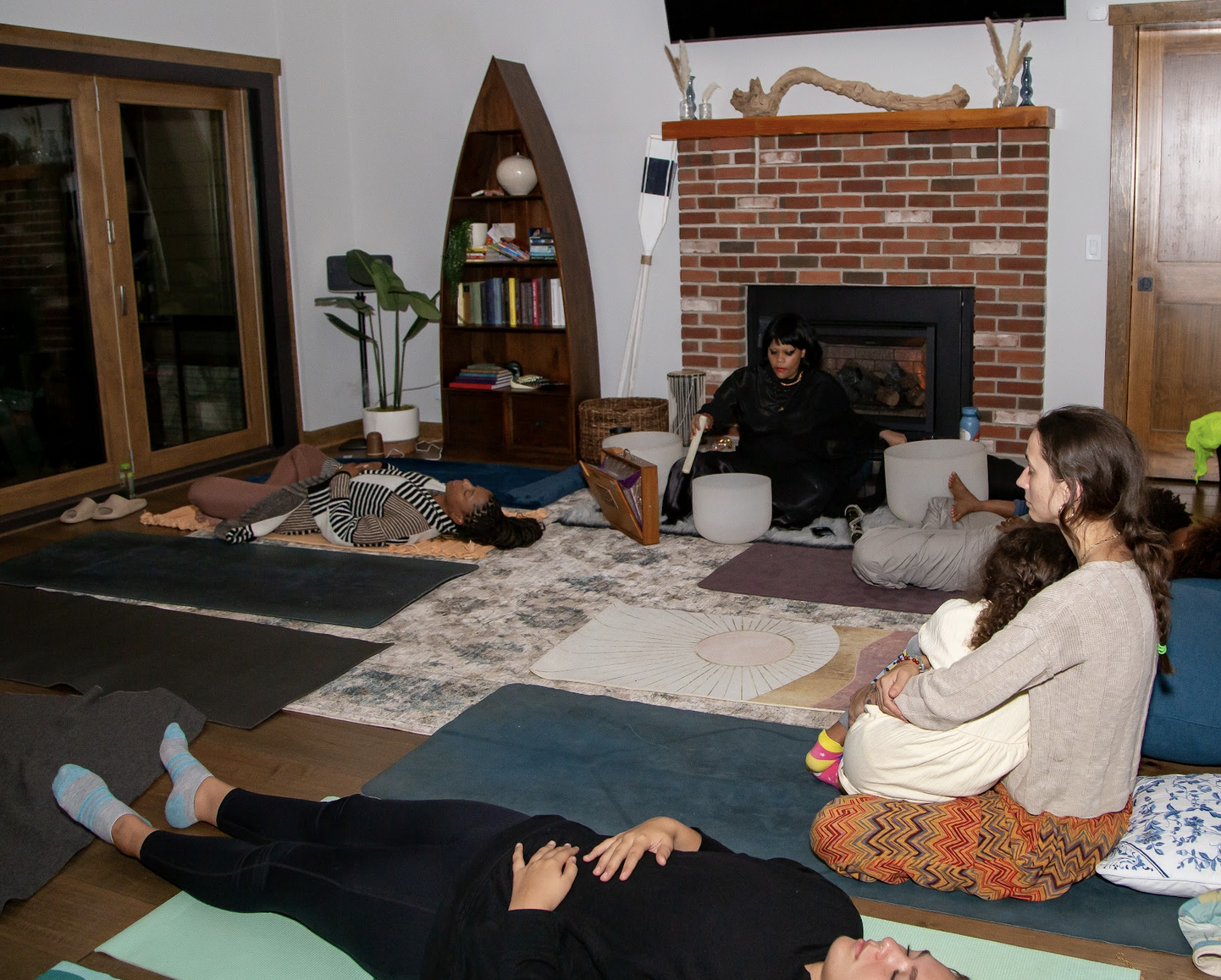 A group of people participating in a meditation or sound healing session in a cozy living room with a brick fireplace, some people lying on yoga mats and others sitting cross-legged. One woman is playing crystal bowls.