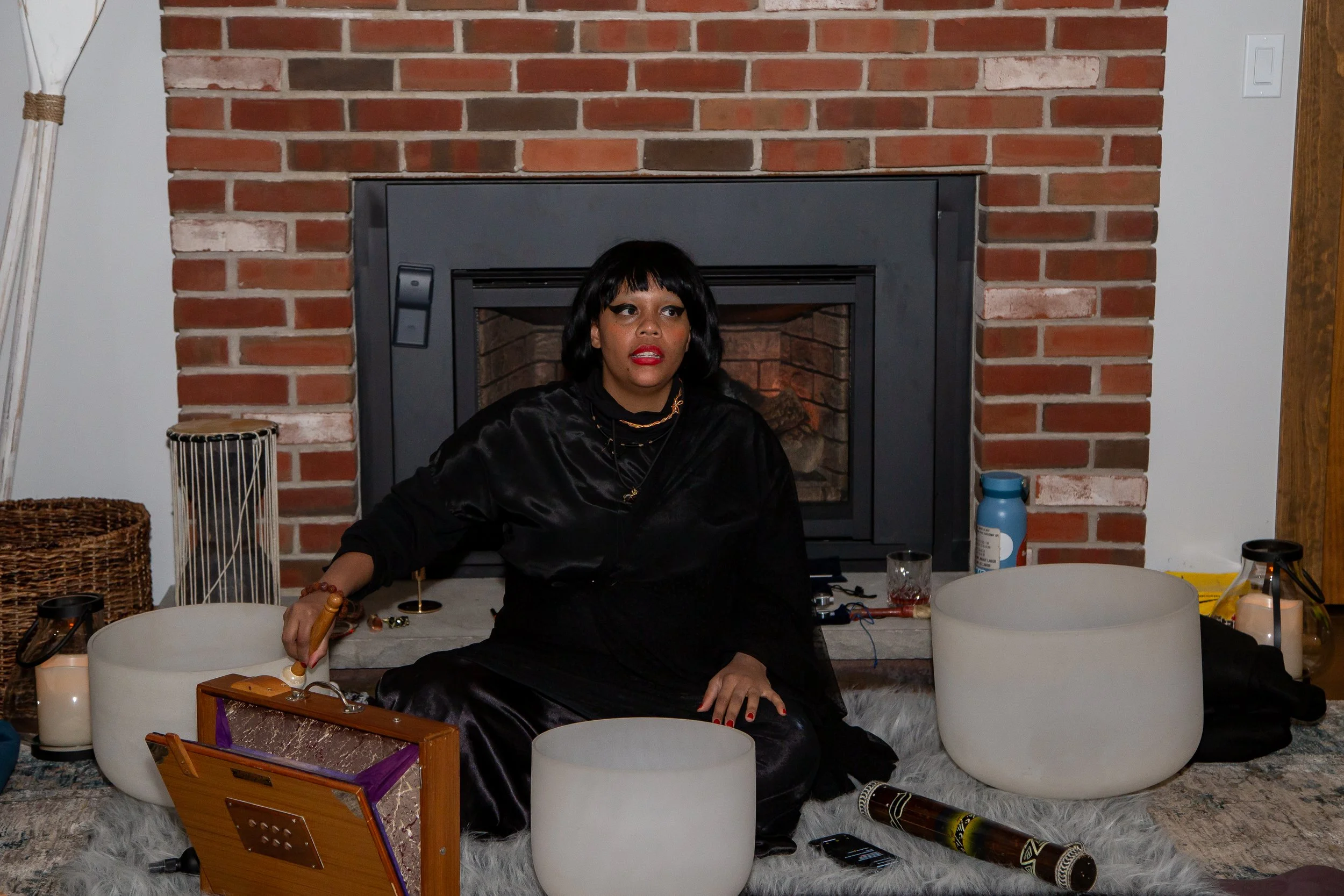 Woman sitting on a rug in front of a brick fireplace, surrounded by white singing bowls, a wooden box, candles, and various objects.