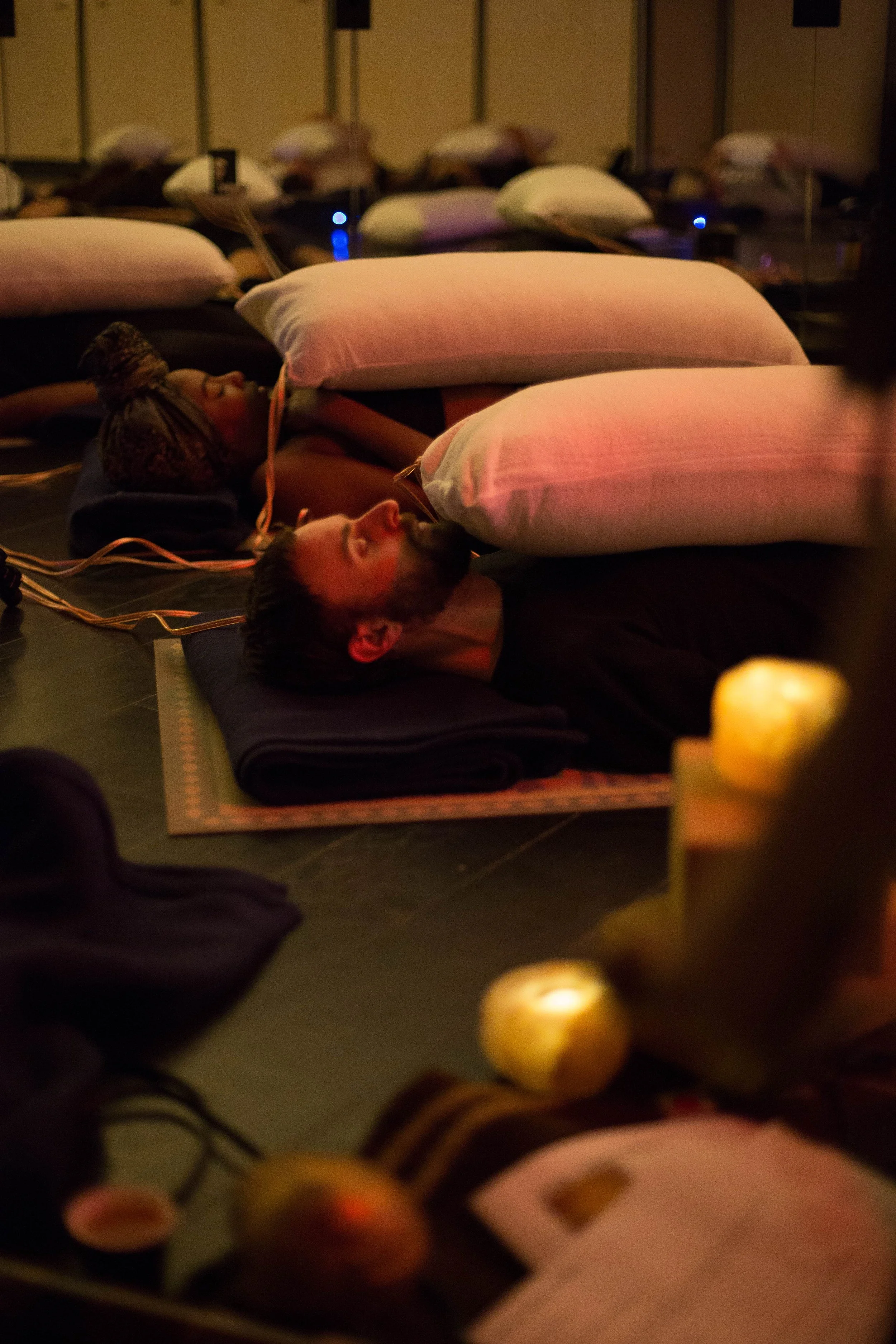 People lying down on mats with pillows, participating in a meditation or relaxation session in a dimly lit room with candles.
