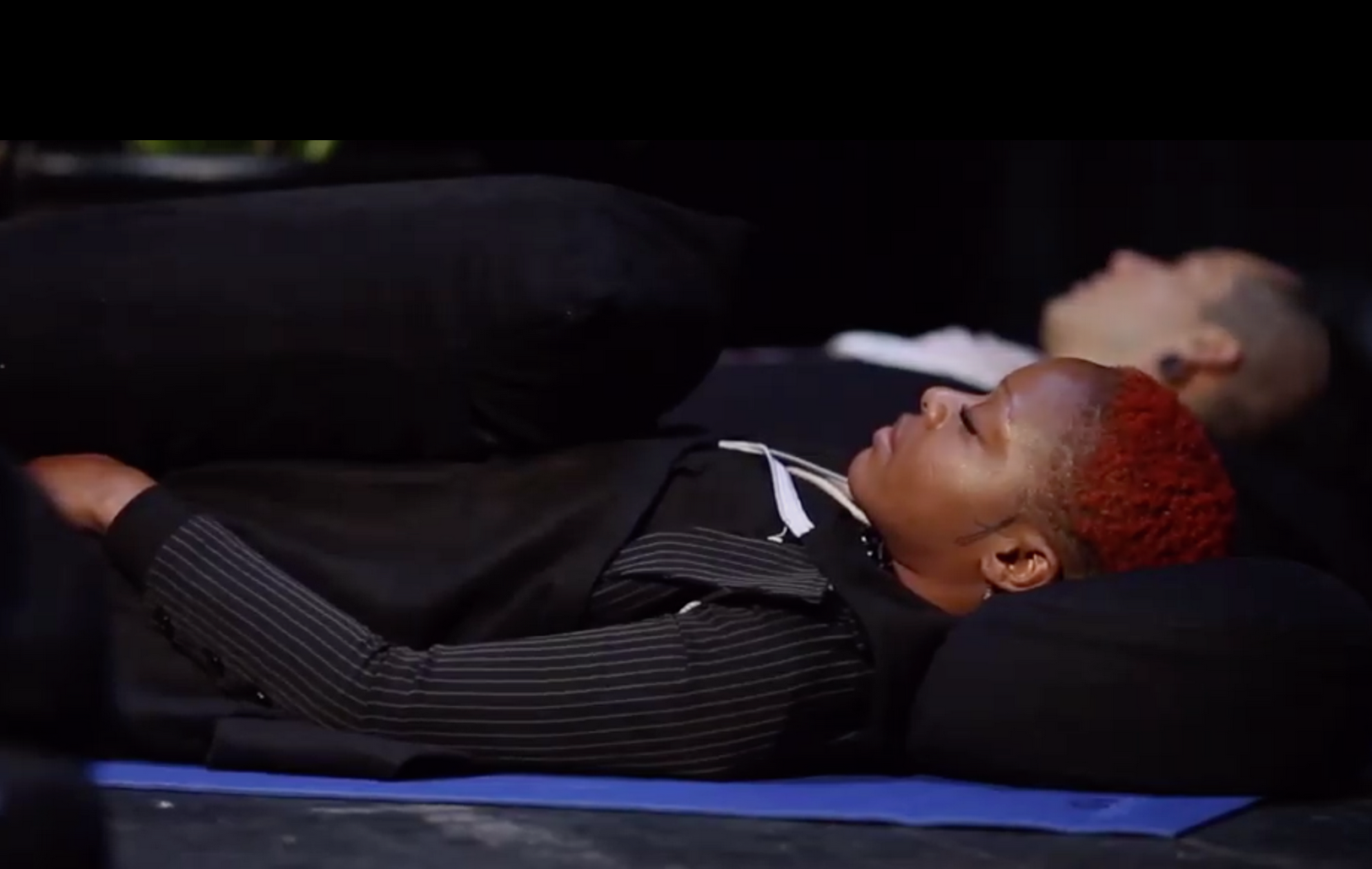 Two individuals sleeping on a black surface, lying on their backs with eyes closed, one with a red beanie and striped long sleeve shirt, the other with light hair and a white shirt, in a dark environment.