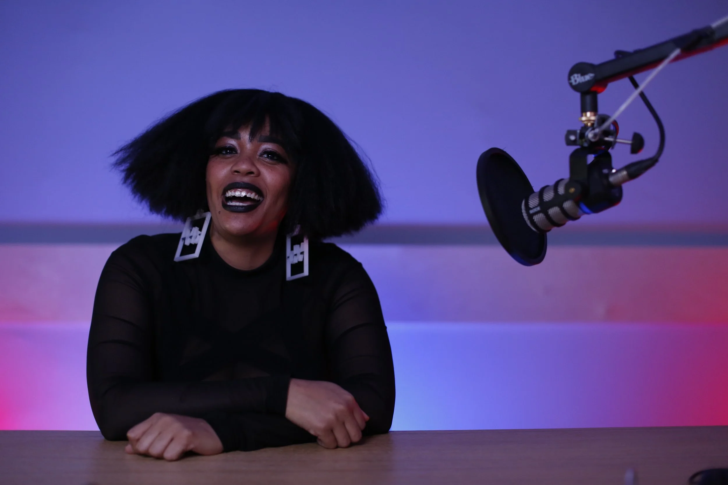 A woman with black hair, dark lipstick, and oversized earrings, smiling and sitting at a table in front of a microphone with colorful purple and pink lighting in the background.