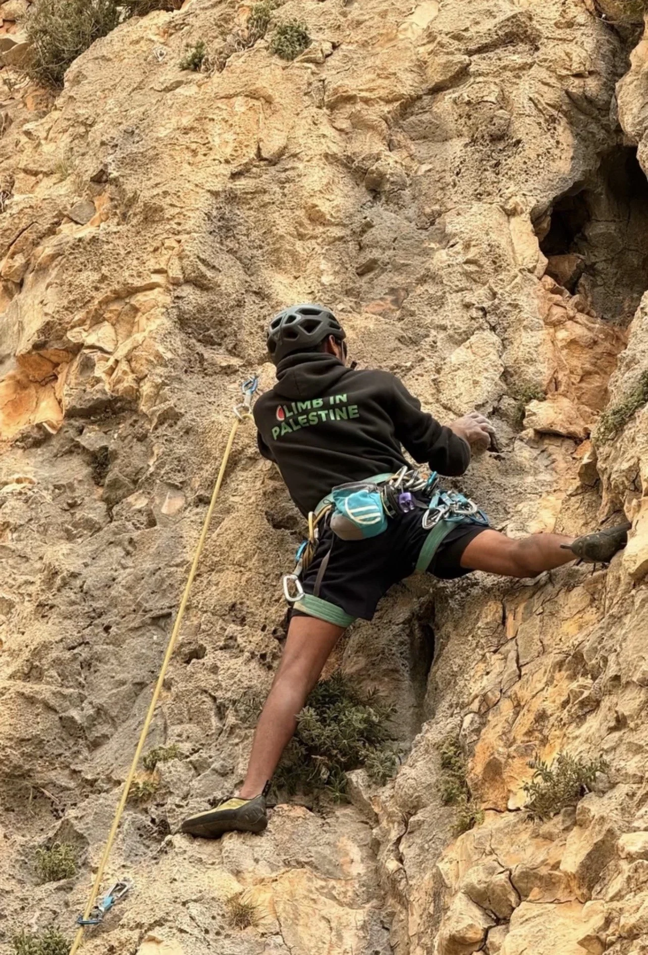 A person rock climbing on a rugged outdoor rocky cliff, wearing a helmet and climbing gear, with a yellow safety rope attached.