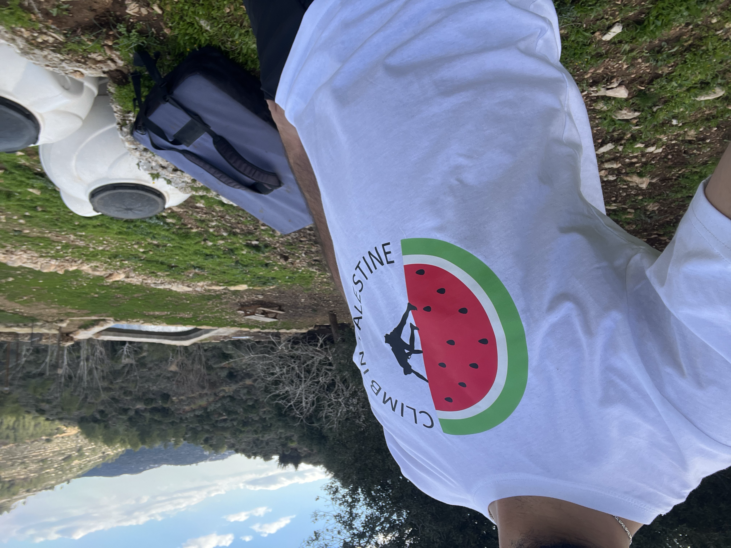 Person lying on the grass in a park during daytime, wearing a white T-shirt with a watermelon design and carrying a black backpack. The background shows trees and a cloudy sky.