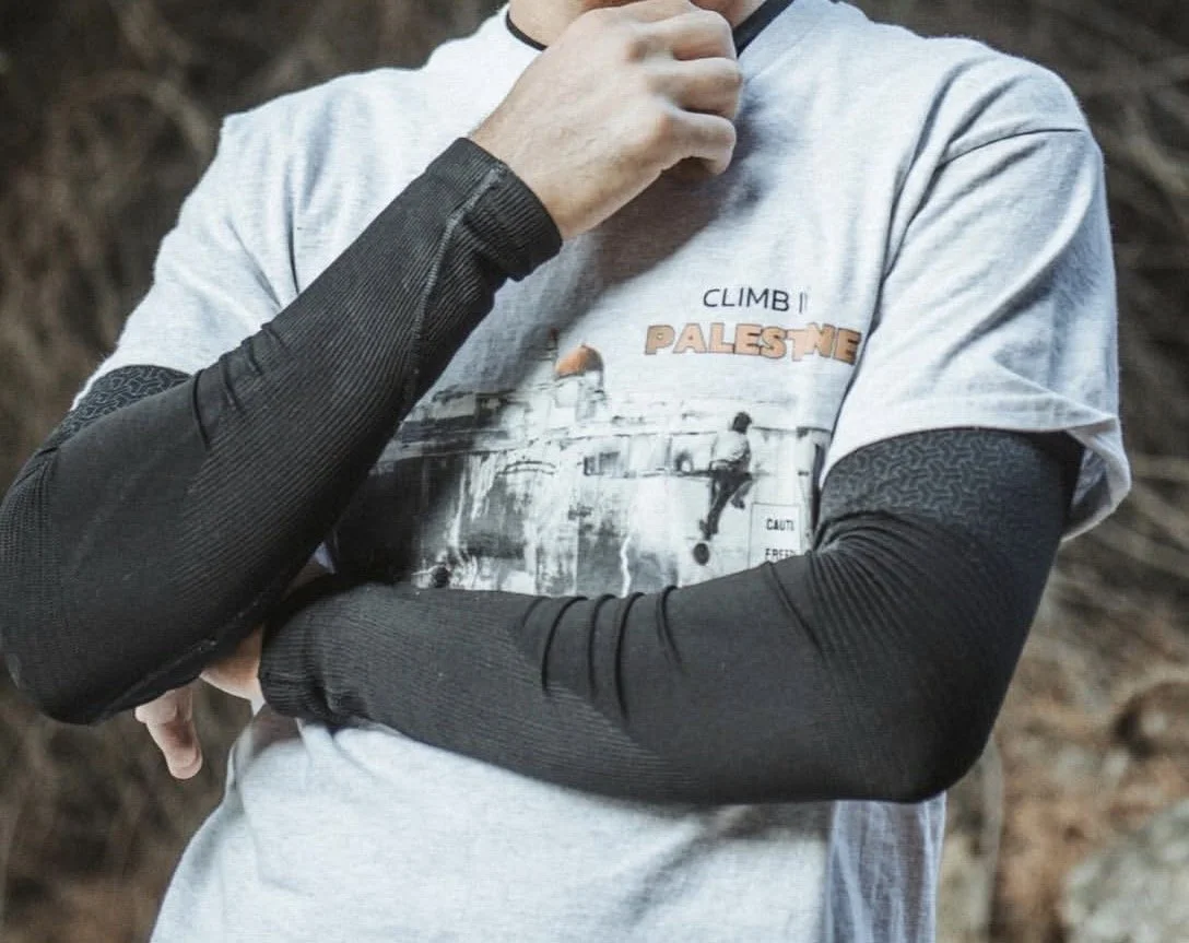 A person wearing a gray T-shirt layered with black arm sleeves, with parts of a graphic design and text that says 'CLIMB PALESTINE' visible on the shirt.