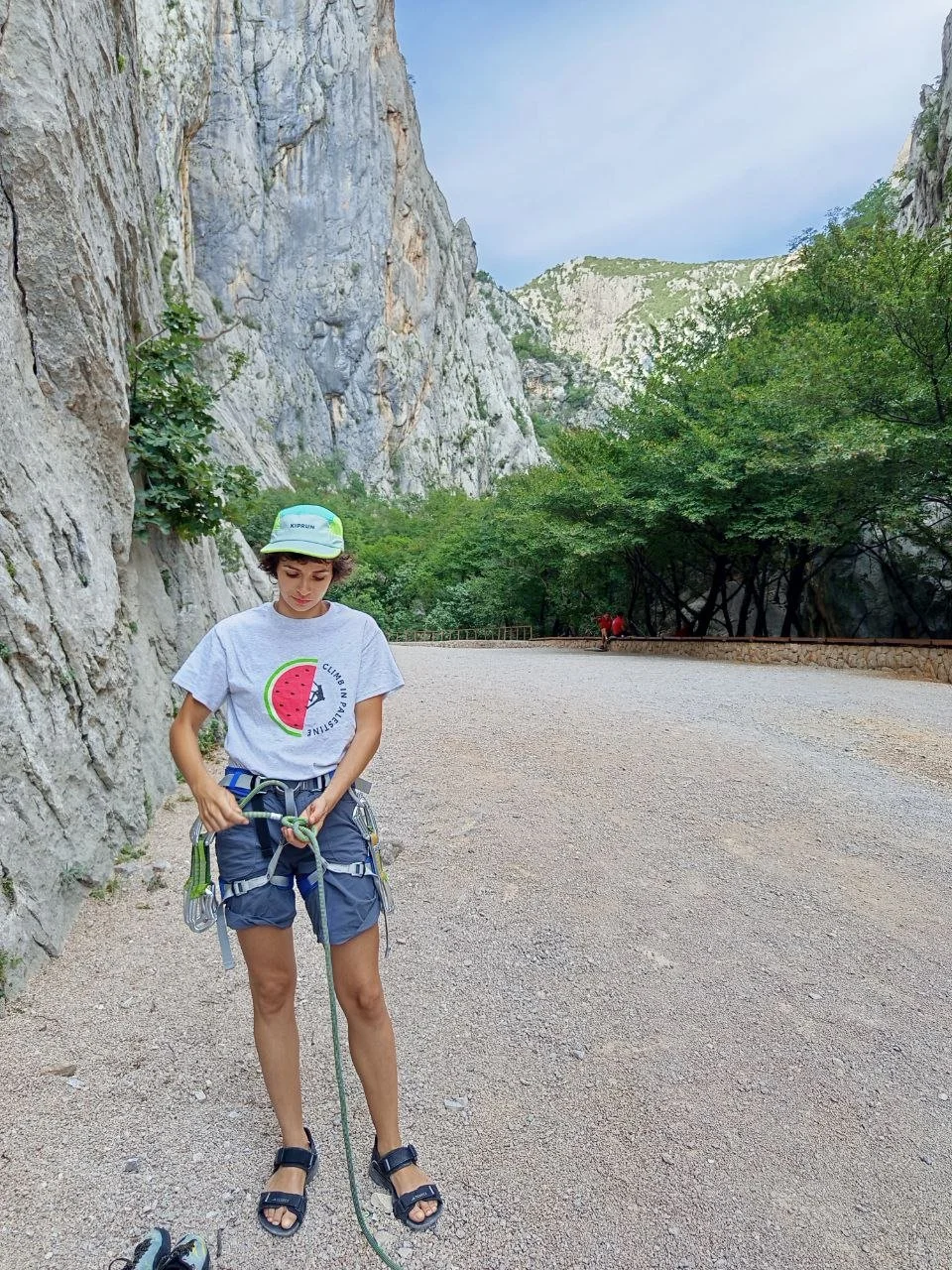 A young person with curly hair wearing a cap, a graphic t-shirt, and shorts stands on a gravel trail, adjusting a climbing harness and rope, with rocky cliffs and green trees in the background.