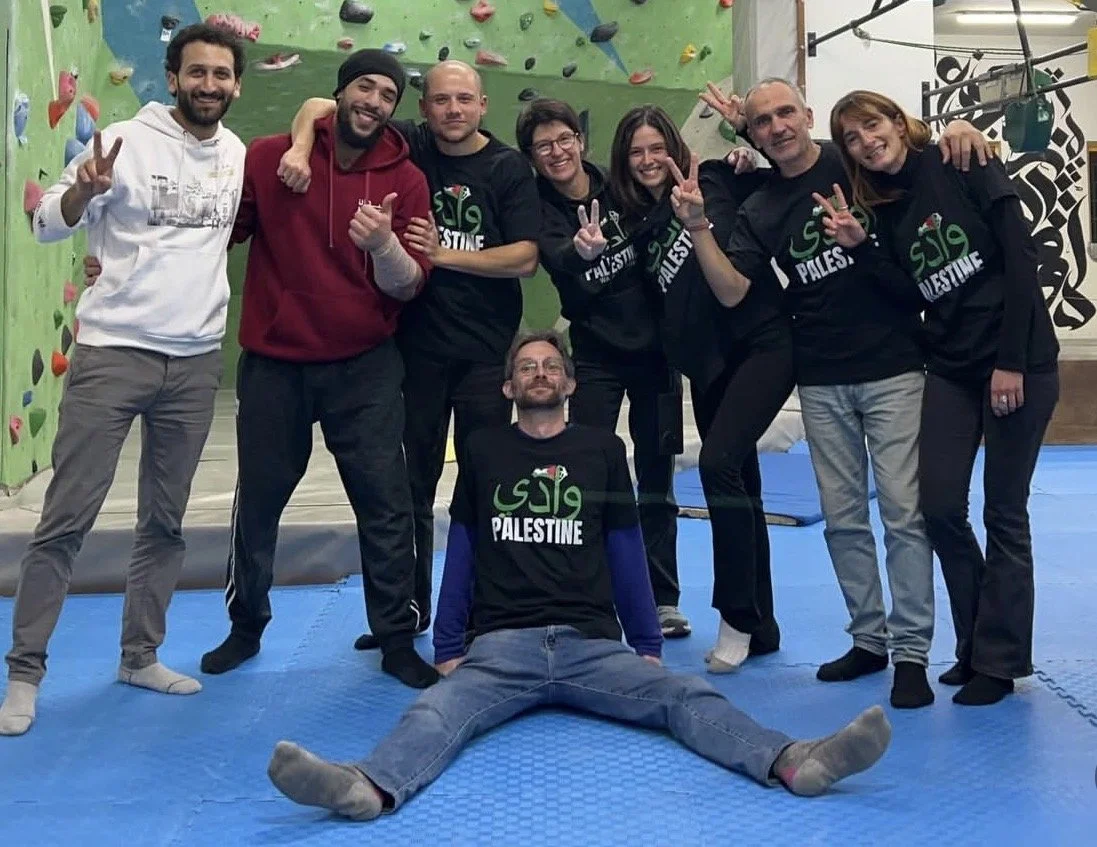 Group of eight people at an indoor rock climbing gym, smiling and making peace signs, with one person sitting on the floor in front doing a split. Some are wearing black shirts with 'Palestine' written on them.