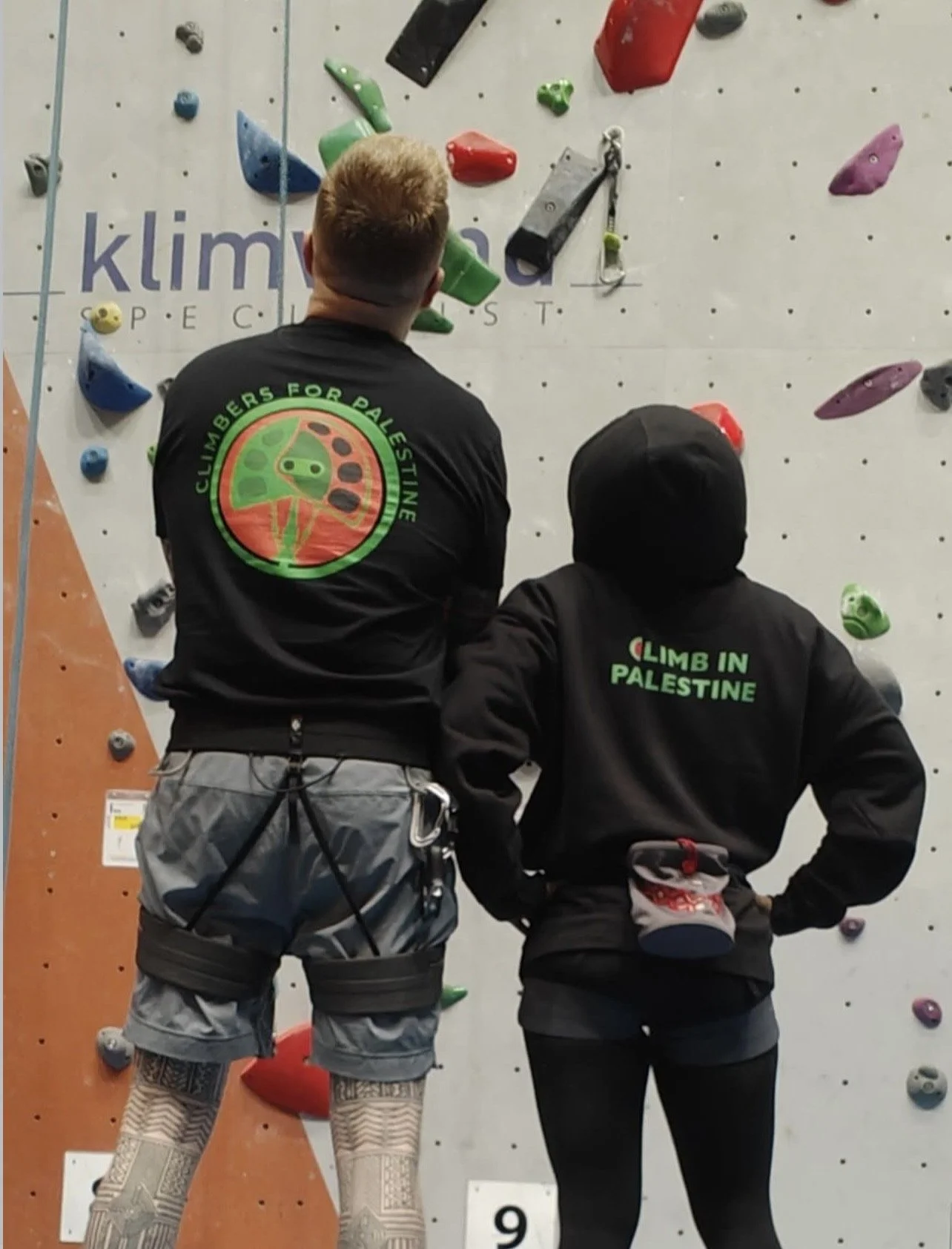 Two climbers facing a climbing wall with colored holds, one wearing a shirt with an apple logo and the words "Climbers for Palestine," and the other wearing a hoodie with the words "Climb in Palestine".
