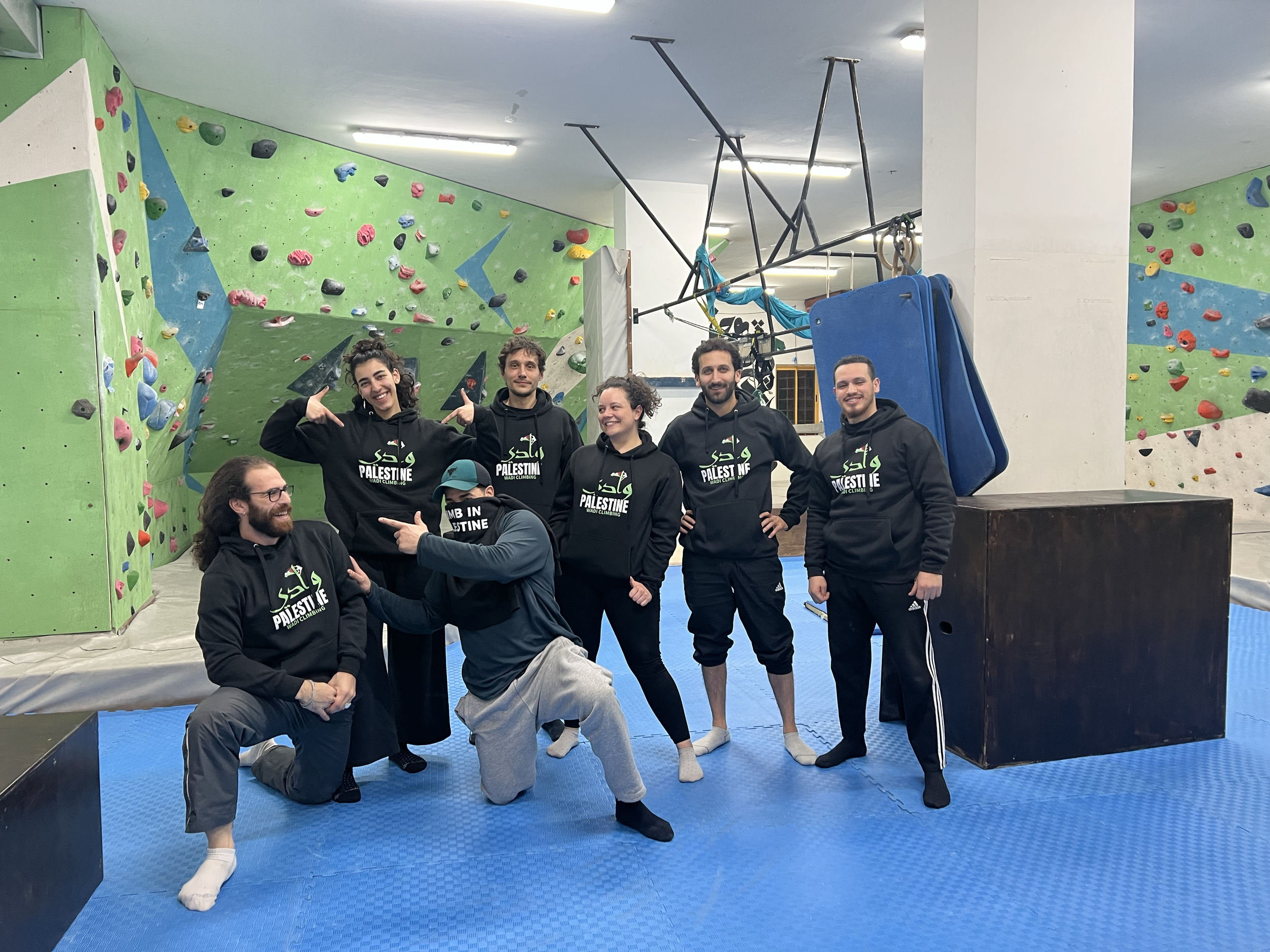 Group of five people in black hoodies with 'Palestine' written on them, posing in an indoor climbing gym with colorful climbing walls and blue padded flooring.