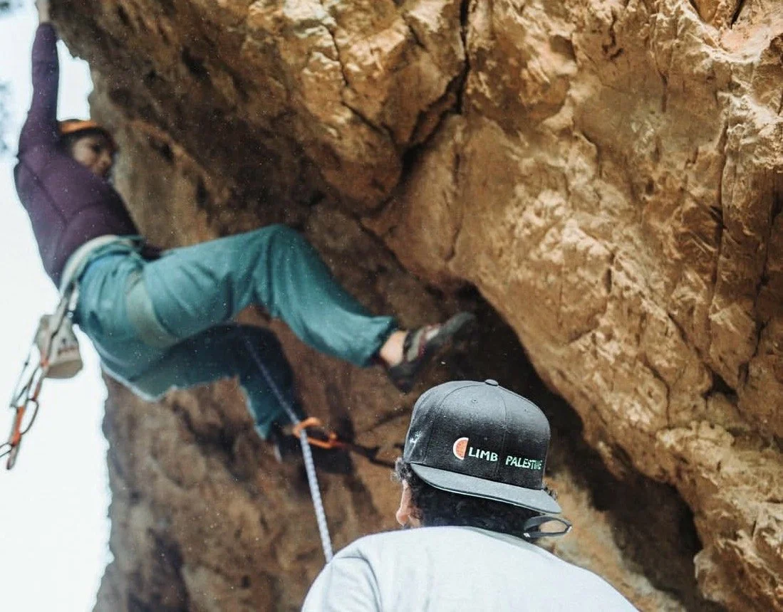 A person in a purple hoodie and teal pants climbing a rock wall outdoors, with a harness and rope, while another person wearing a black cap watches.