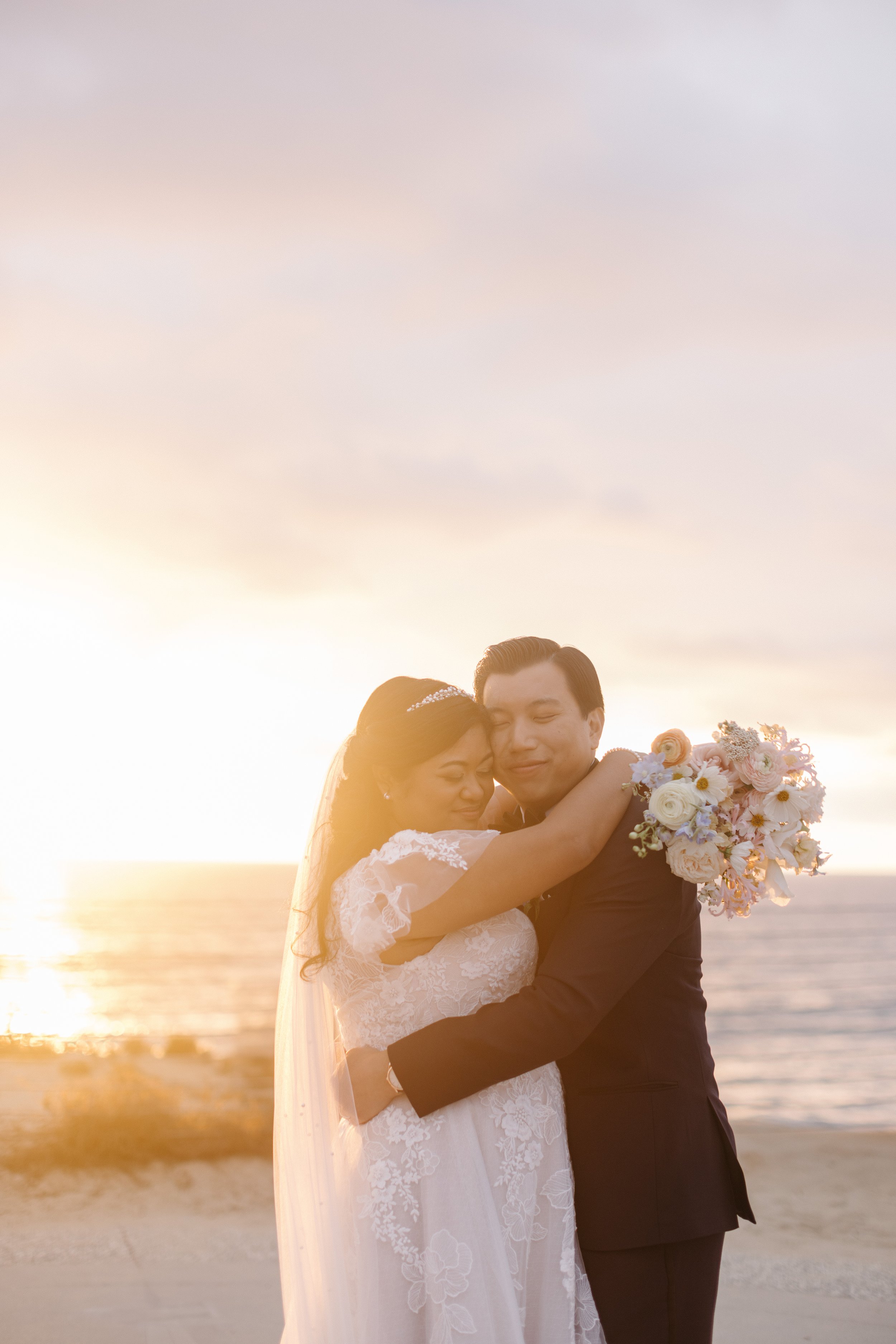 A bride and groom embrace on the beach at sunset, with the ocean and sky in the background, the bride holding a bouquet of white and peach flowers.