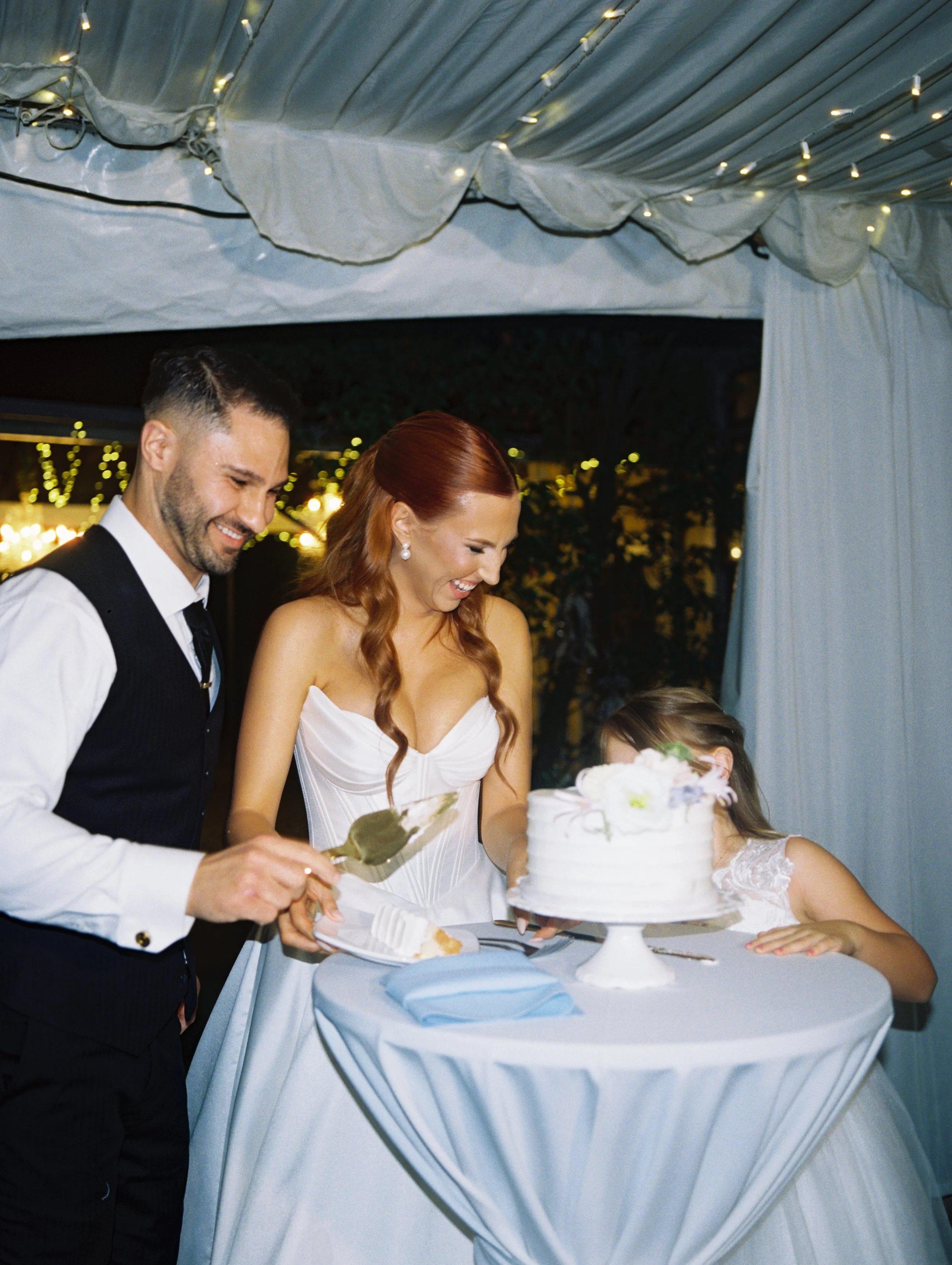 Three people at a wedding reception, a man, a woman in a white gown, and a young girl, are laughing and cutting a cake under a decorated canopy with string lights.
