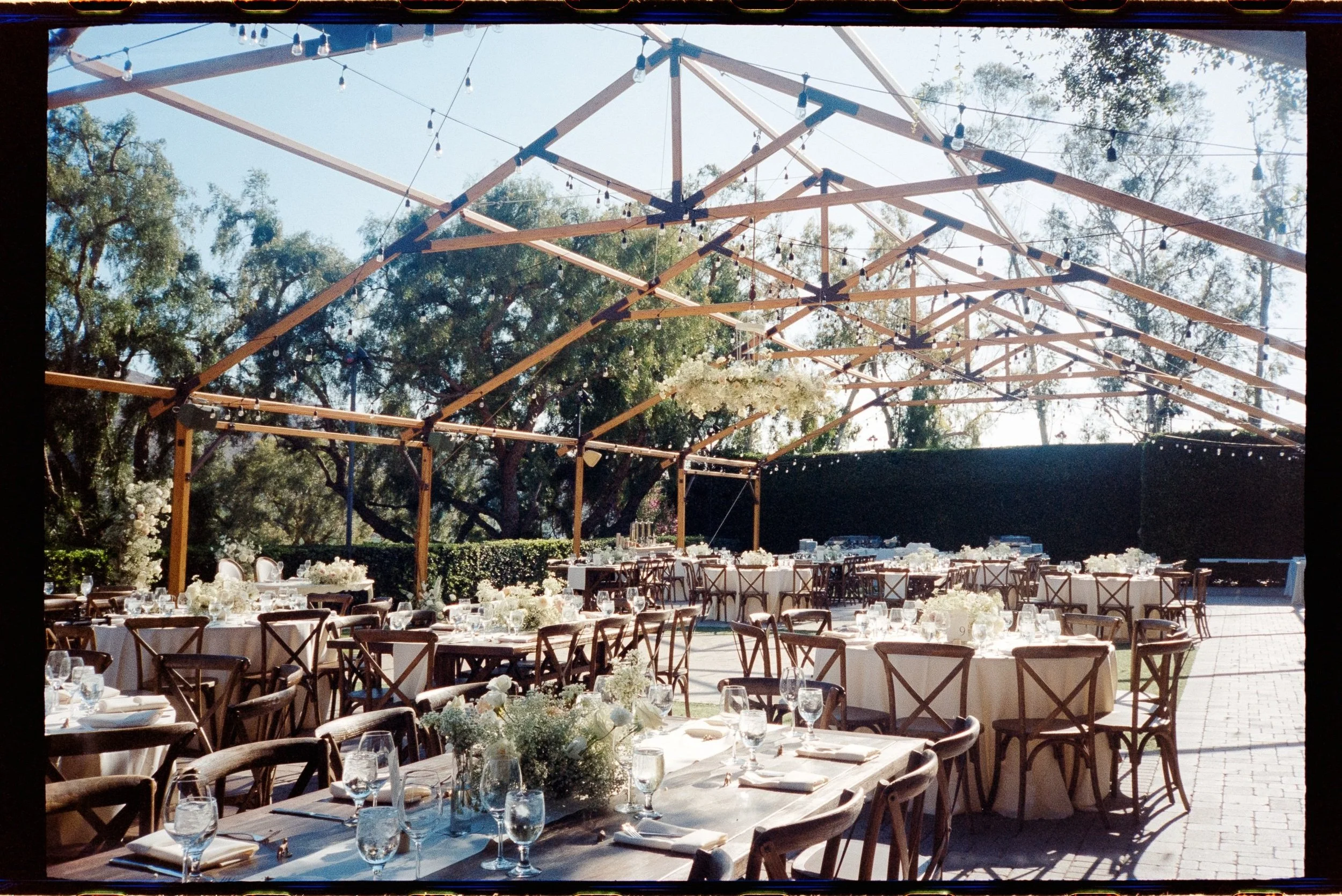 Outdoor event setup with round and rectangular tables, decorated with white flowers, glassware, and tableware, under a wooden geometric frame with string lights, in a garden setting.