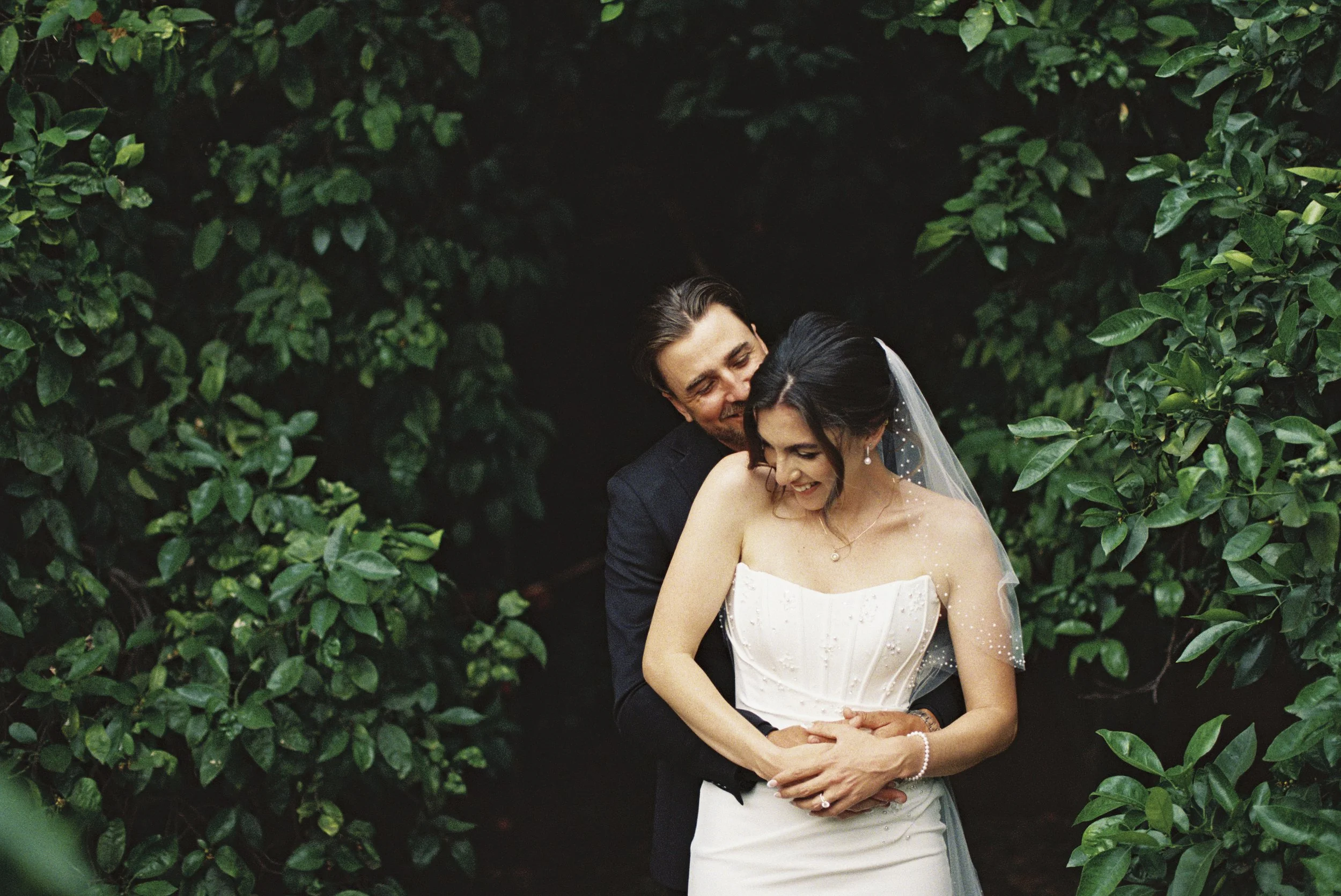 Bride and groom embracing among lush greenery in a romantic wedding portrait photographed on film.