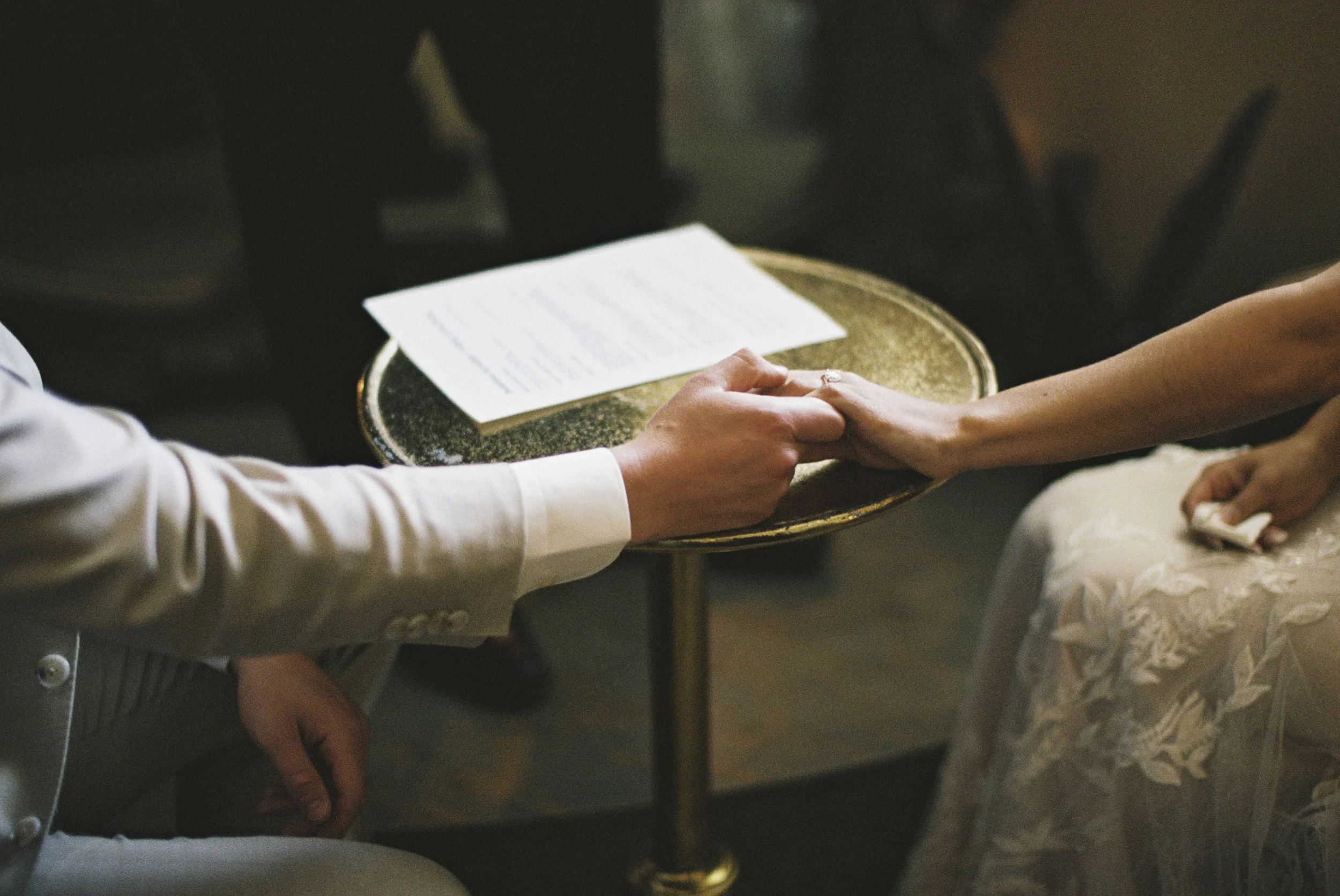 A person holding another person's hand during a wedding ceremony, with a document on a small round table in front of them.