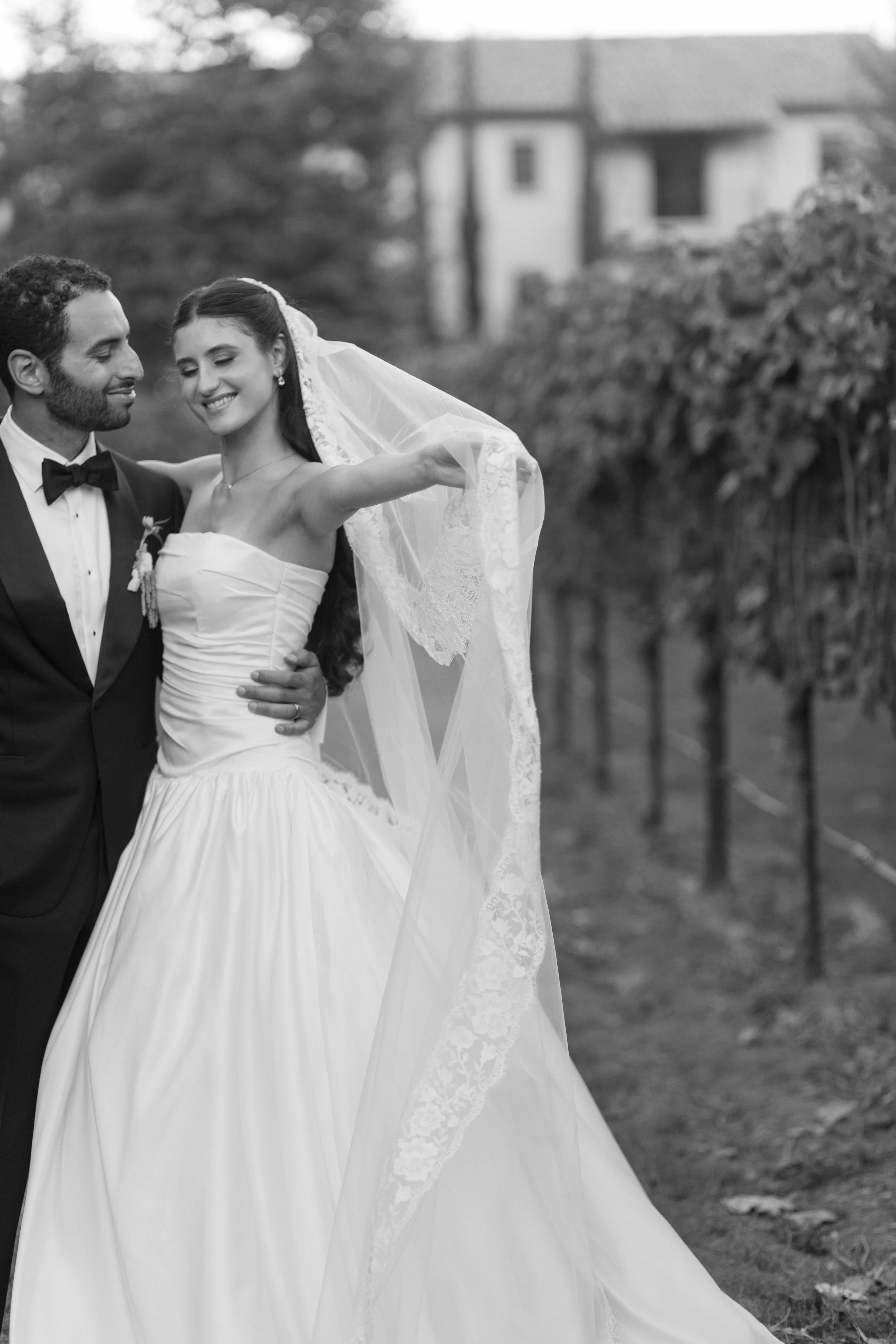 A black and white photo of a newlywed couple outdoors, with the groom in a tuxedo and the bride in a wedding gown, smiling and holding each other.