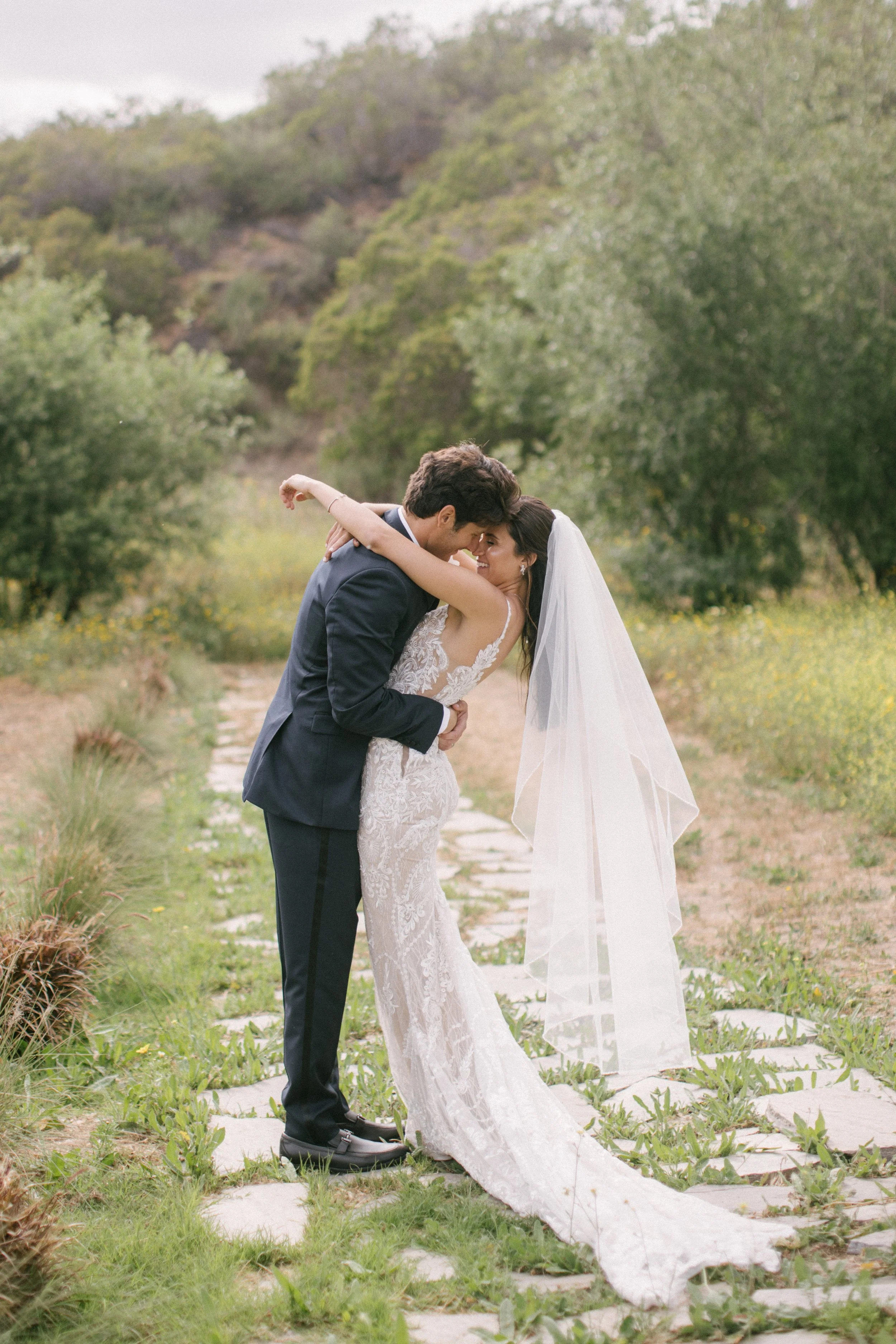 A bride and groom sharing a romantic moment on a scenic outdoor pathway surrounded by greenery.