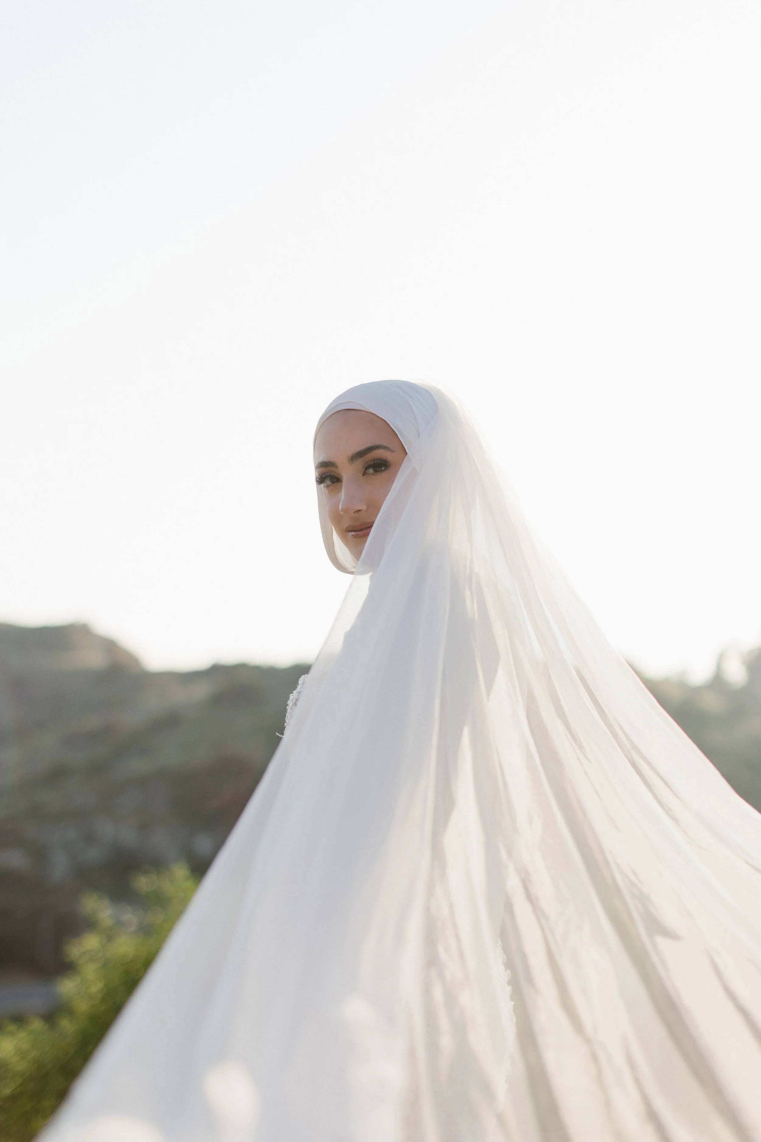 A woman in a white hijab standing outdoors during daylight, with a blurred background of hills and trees.
