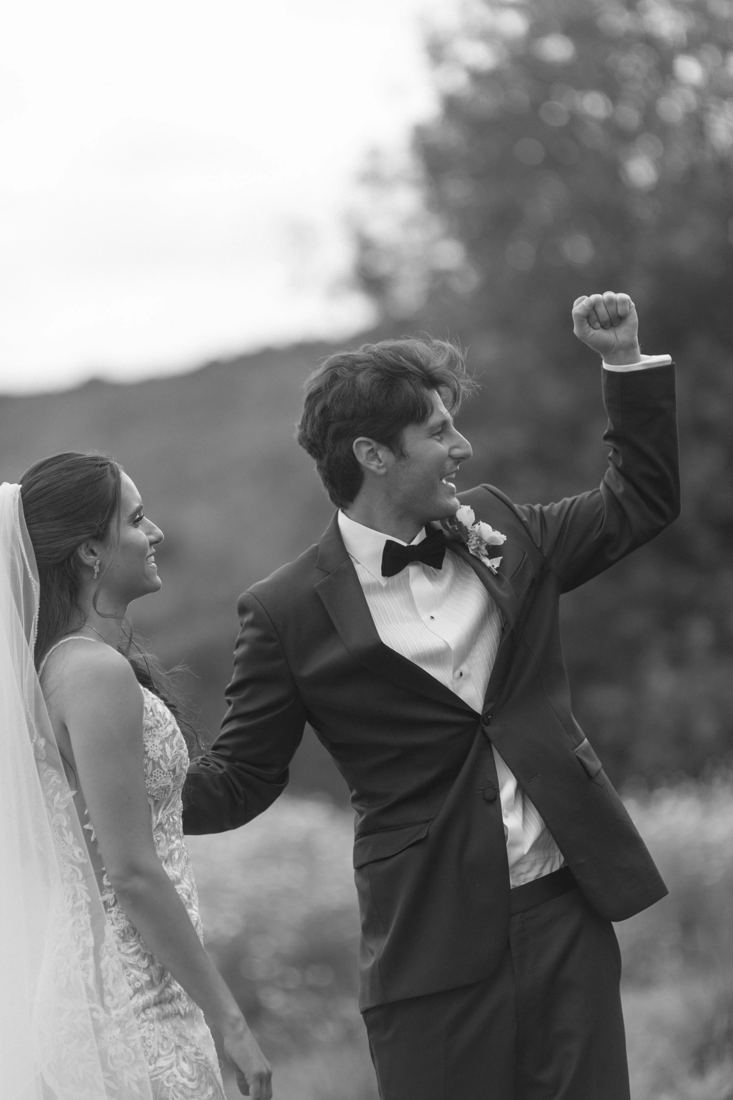 A black and white photo of a newlywed couple outdoors, with the groom raising his arm in celebration and the bride smiling at him, both dressed in wedding attire.