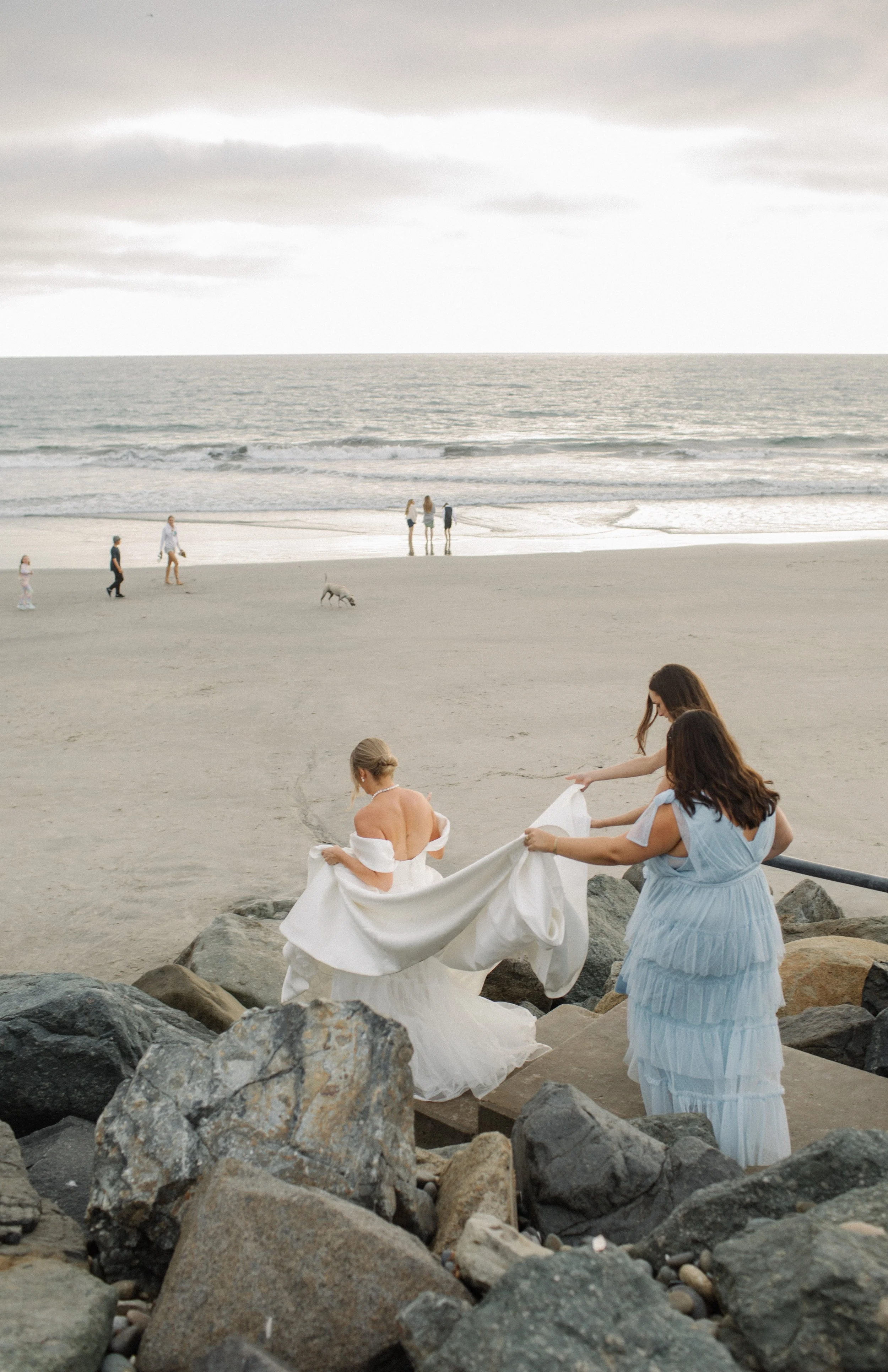 Women preparing for a beach wedding, with a bride in a white wedding dress and two women assisting her, on a rocky pathway overlooking the sandy beach and ocean with other people and dogs in the background.