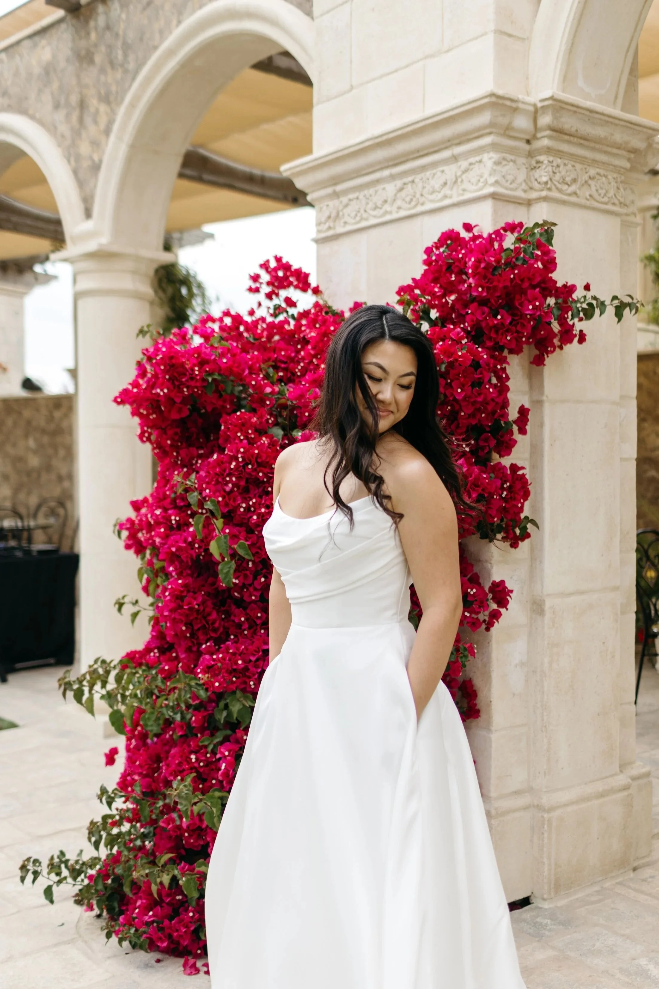A woman in a white wedding dress standing in front of a vibrant pink bougainvillea plant, with stone arches in the background.