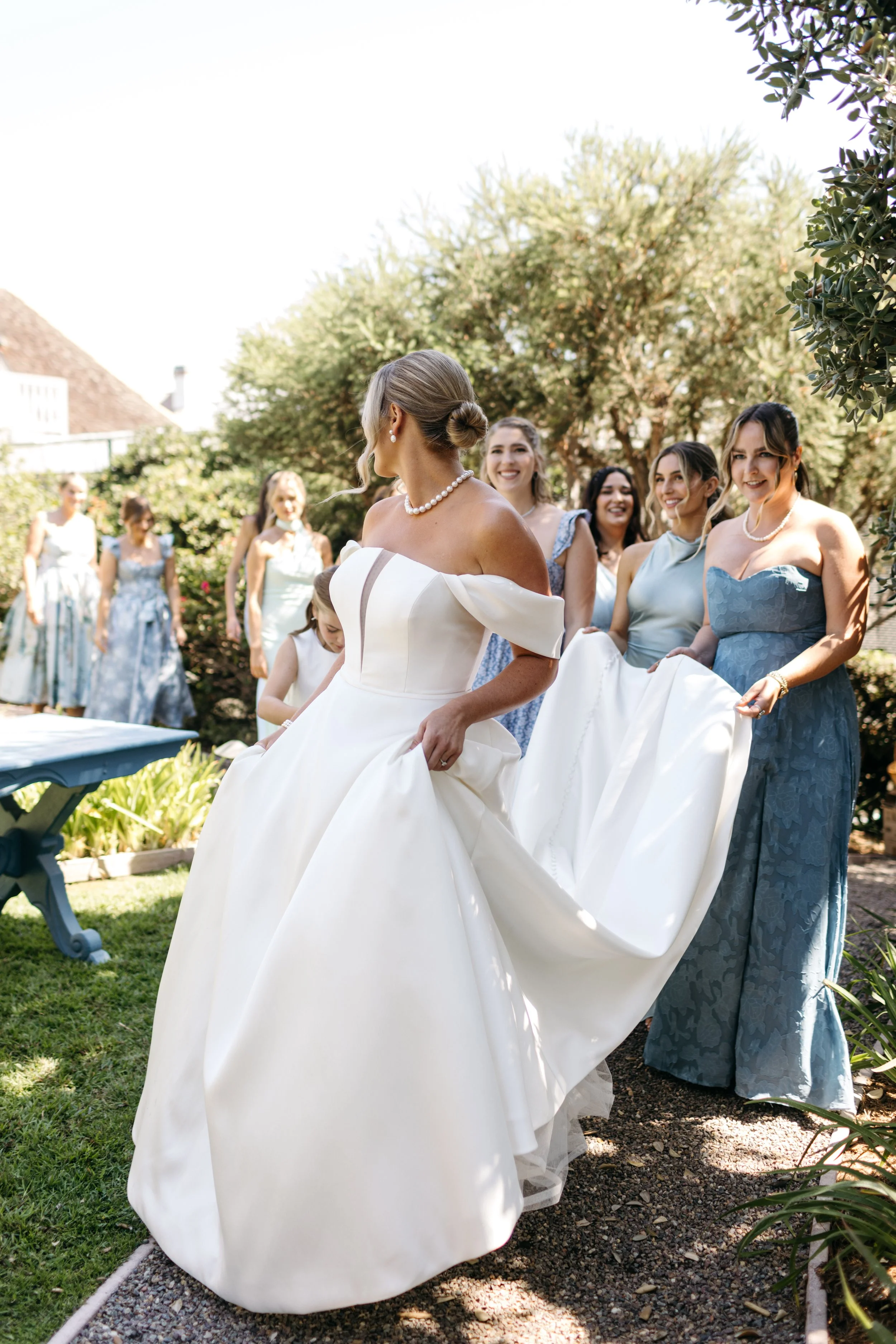 A bride in a white wedding gown with off-shoulder sleeves is outside, surrounded by bridesmaids in blue dresses, during a wedding celebration.