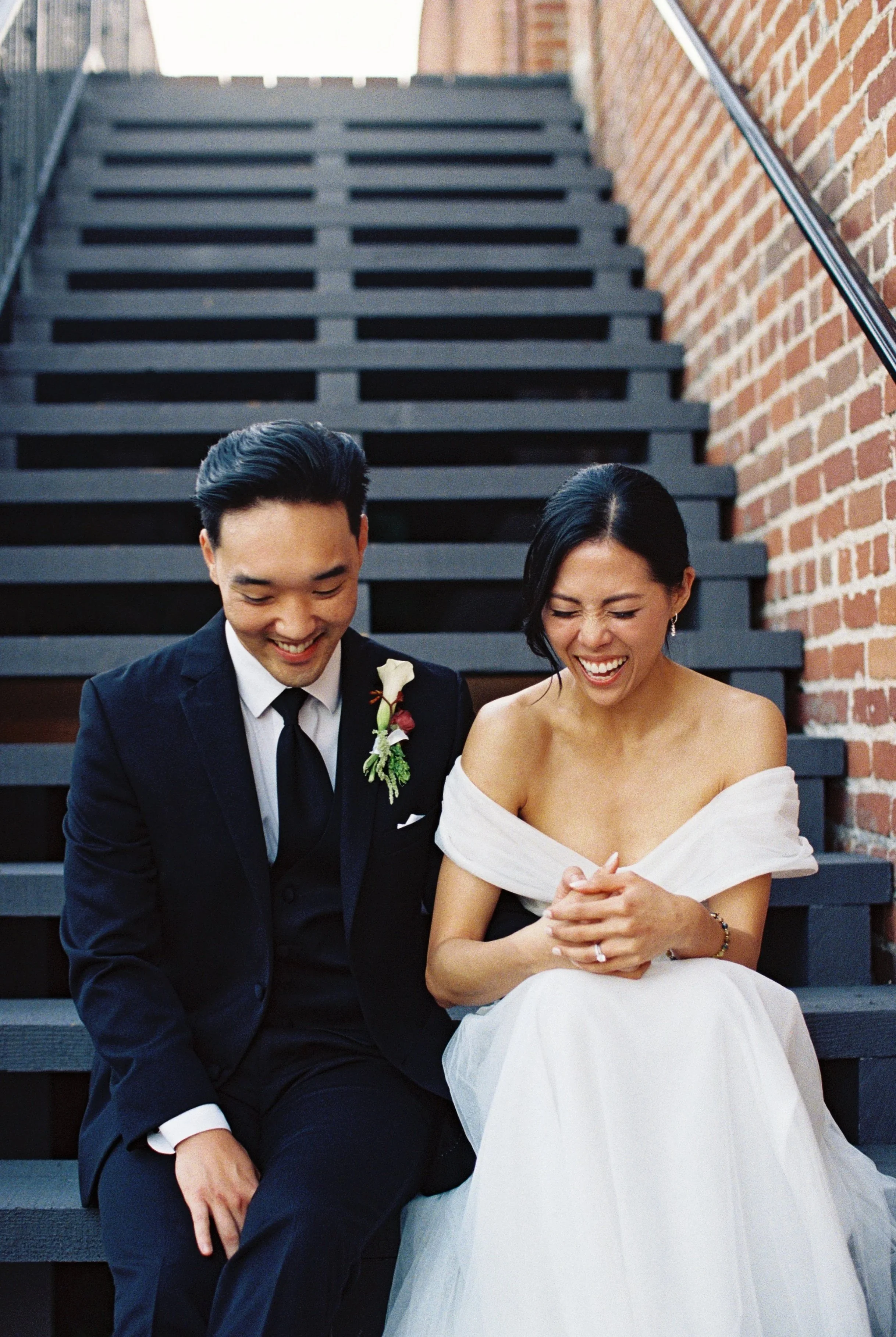 A bride and groom sitting on a staircase, laughing together during a wedding photo shoot.