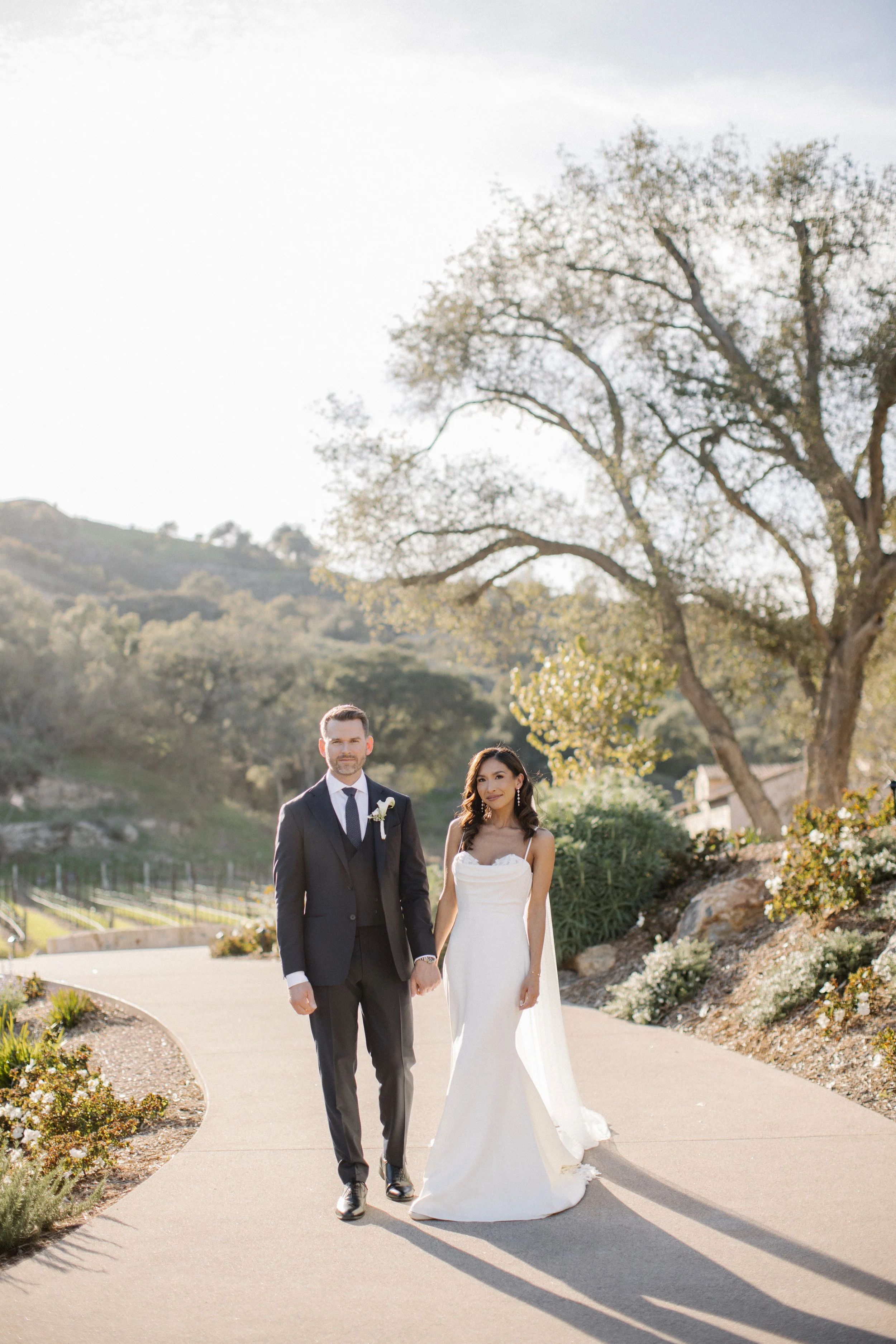 A bride and groom holding hands walking outdoors on a sunny day, surrounded by trees and hills.