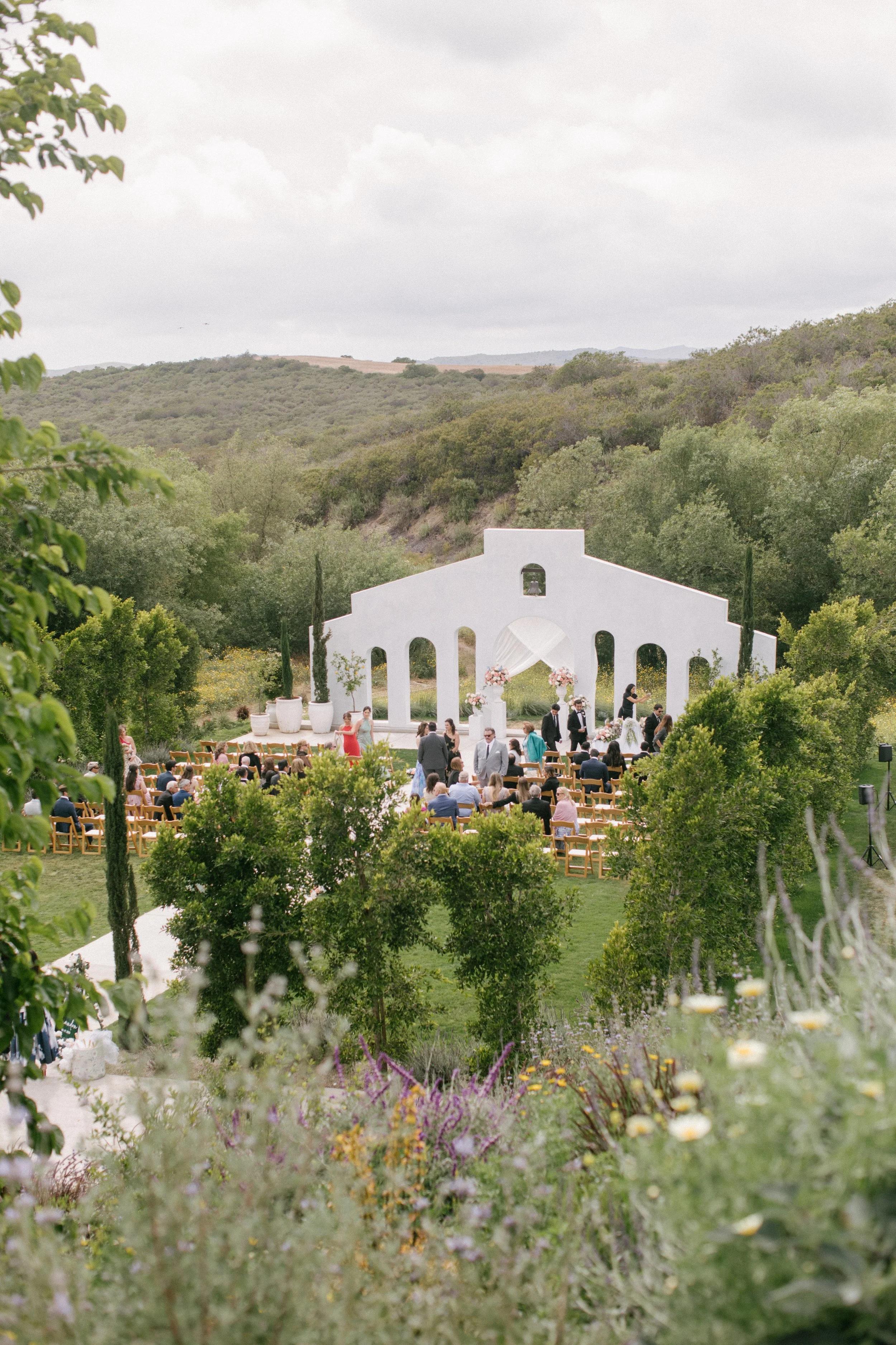 An outdoor wedding ceremony setup in a lush green landscape with a white arch and seated guests.