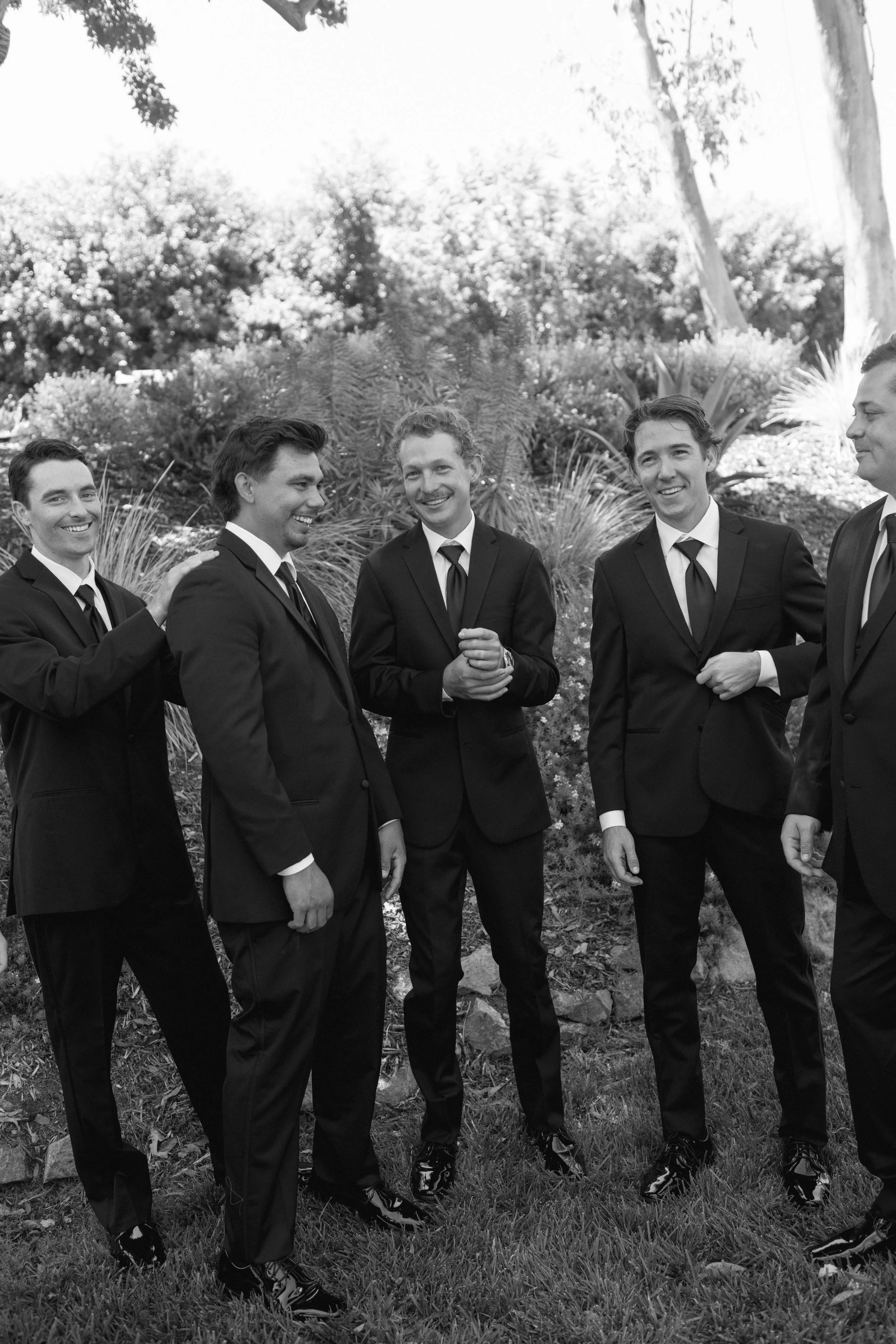 → Groomsmen laughing together outdoors in a candid black-and-white wedding photograph.