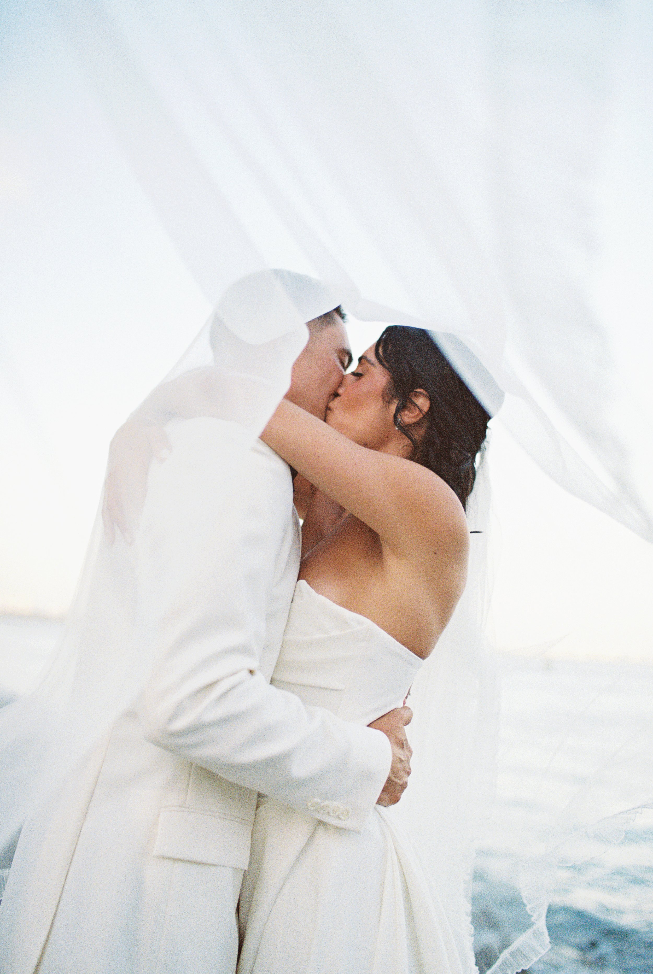 A couple in wedding attire embracing and kissing under a sheer veil, near the water at sunset.