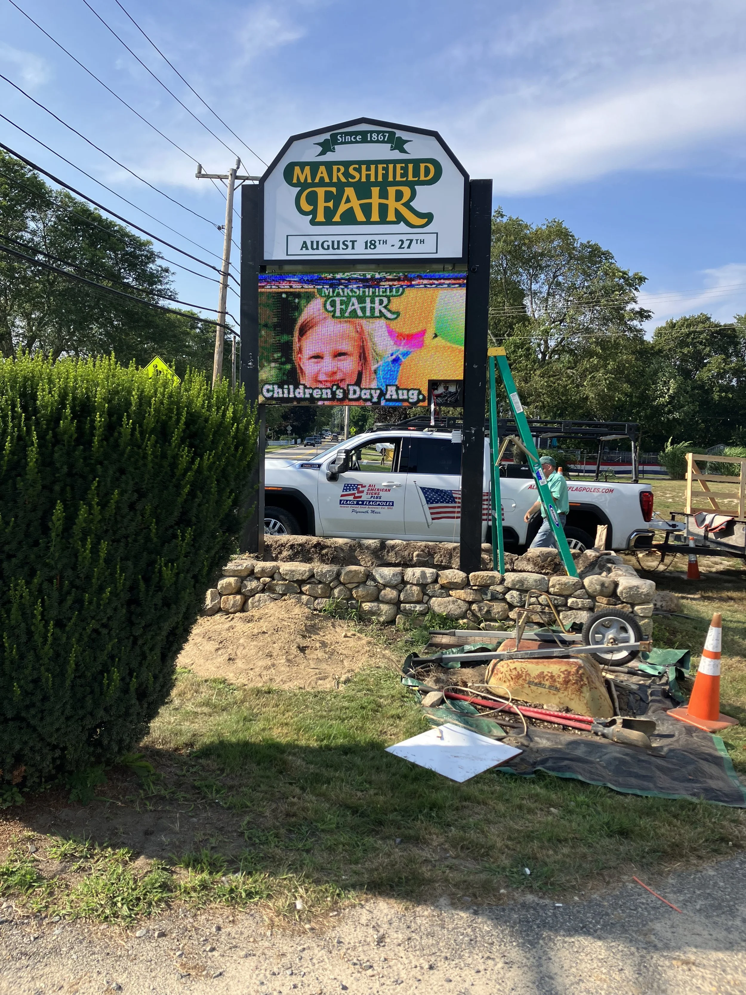 Sign for Marshfield Fair with dates August 18-27, the sign is being installed, with a ladder and work tools nearby, and a truck parked in the background.