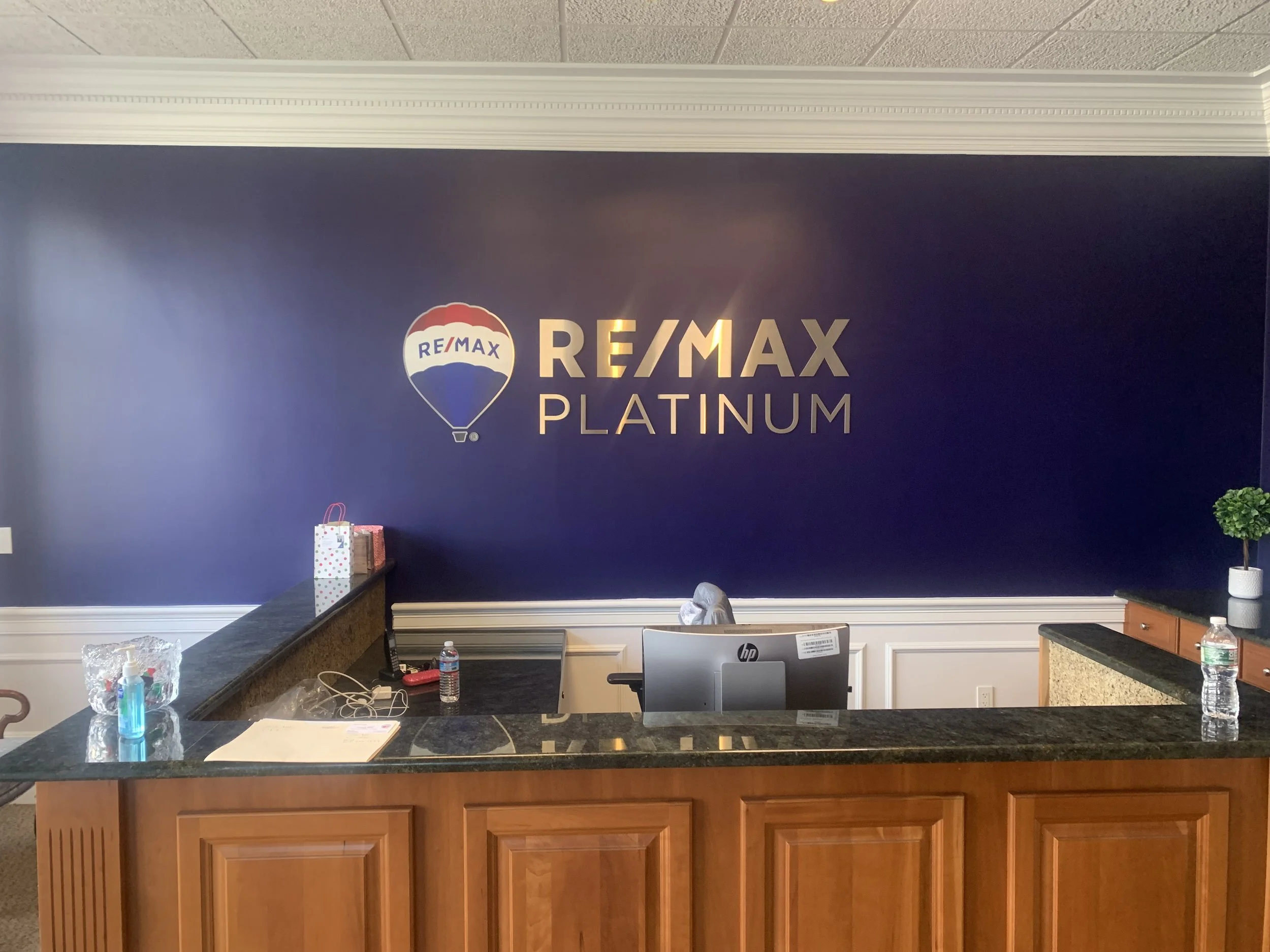 RE/MAX Platinum office reception area with a dark blue wall, a reception desk with bottled water, sanitizer, papers, and a computer, and a potted plant on a side cabinet.