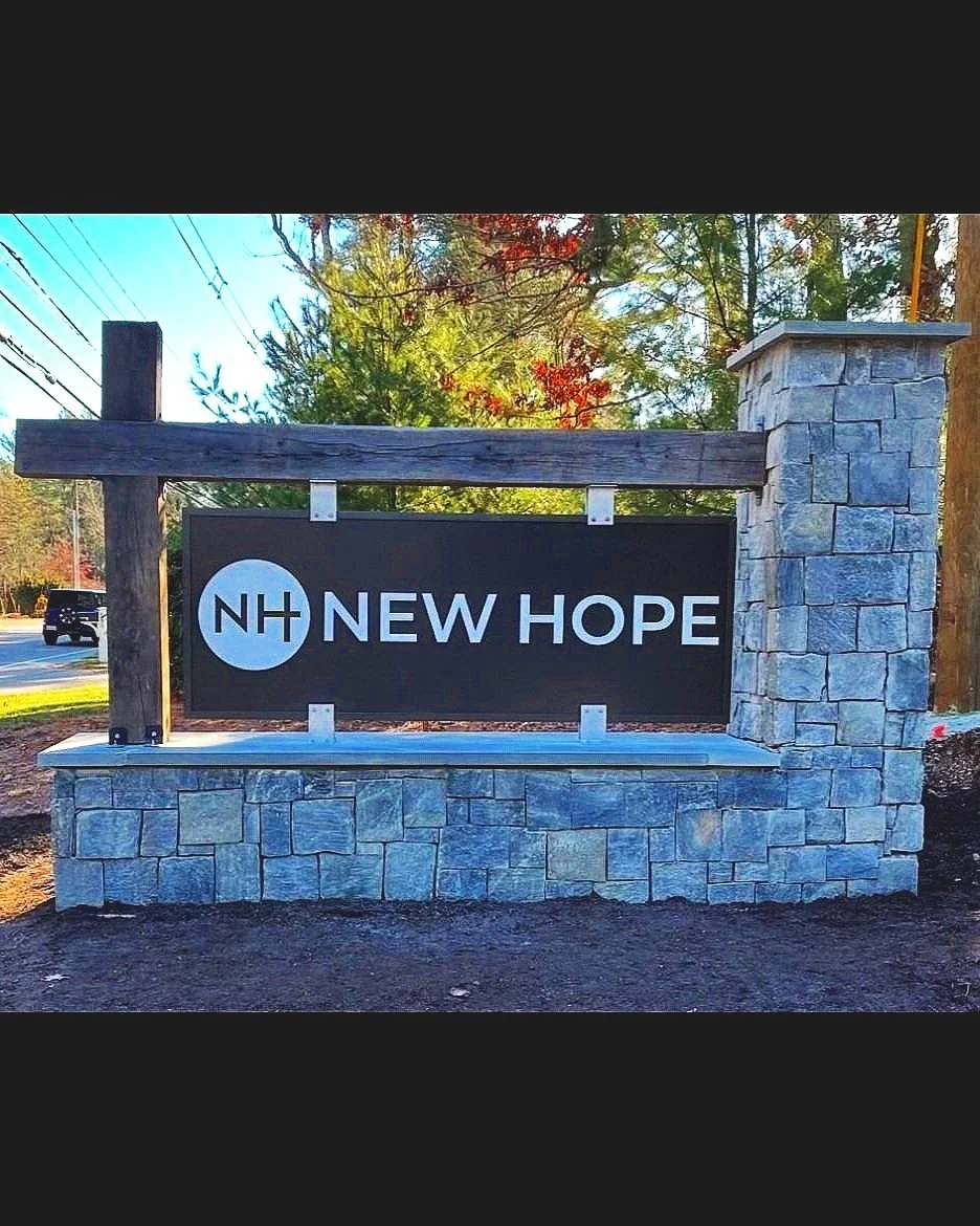Sign on a stone and wood structure reading 'New Hope' with a logo featuring 'NH' in a circle, set outdoors with trees and a road in the background.