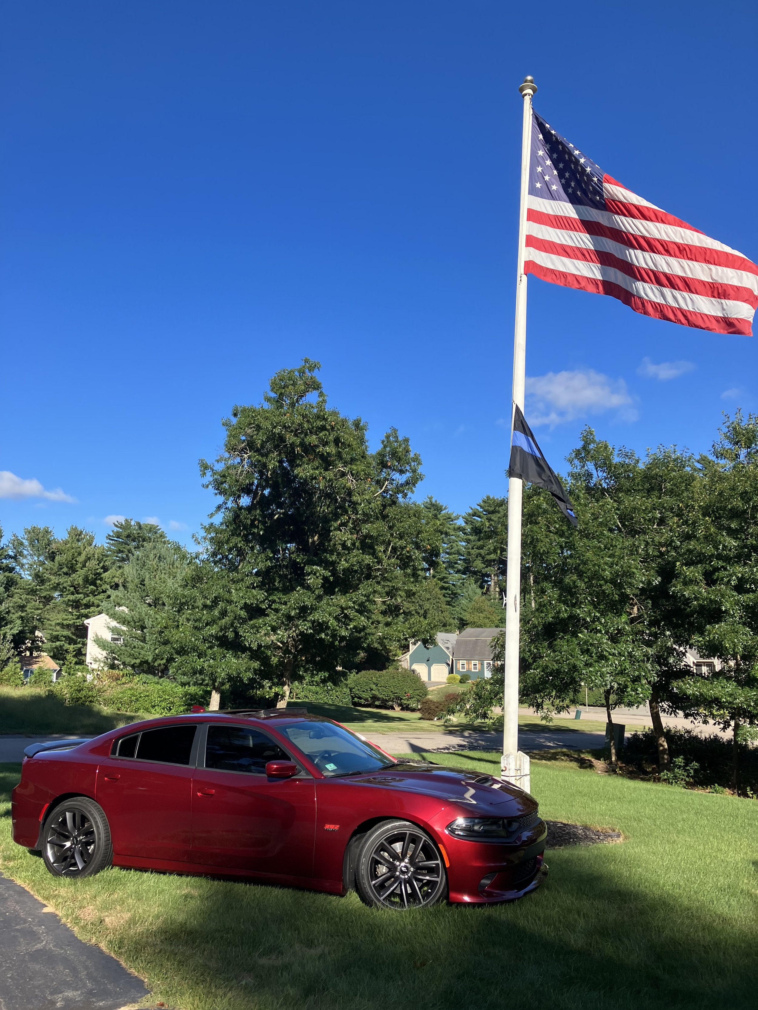A red sports car parked on a grassy area next to a tap with an American flag and a blue flag, under a bright blue sky with some clouds and a background of trees and houses.
