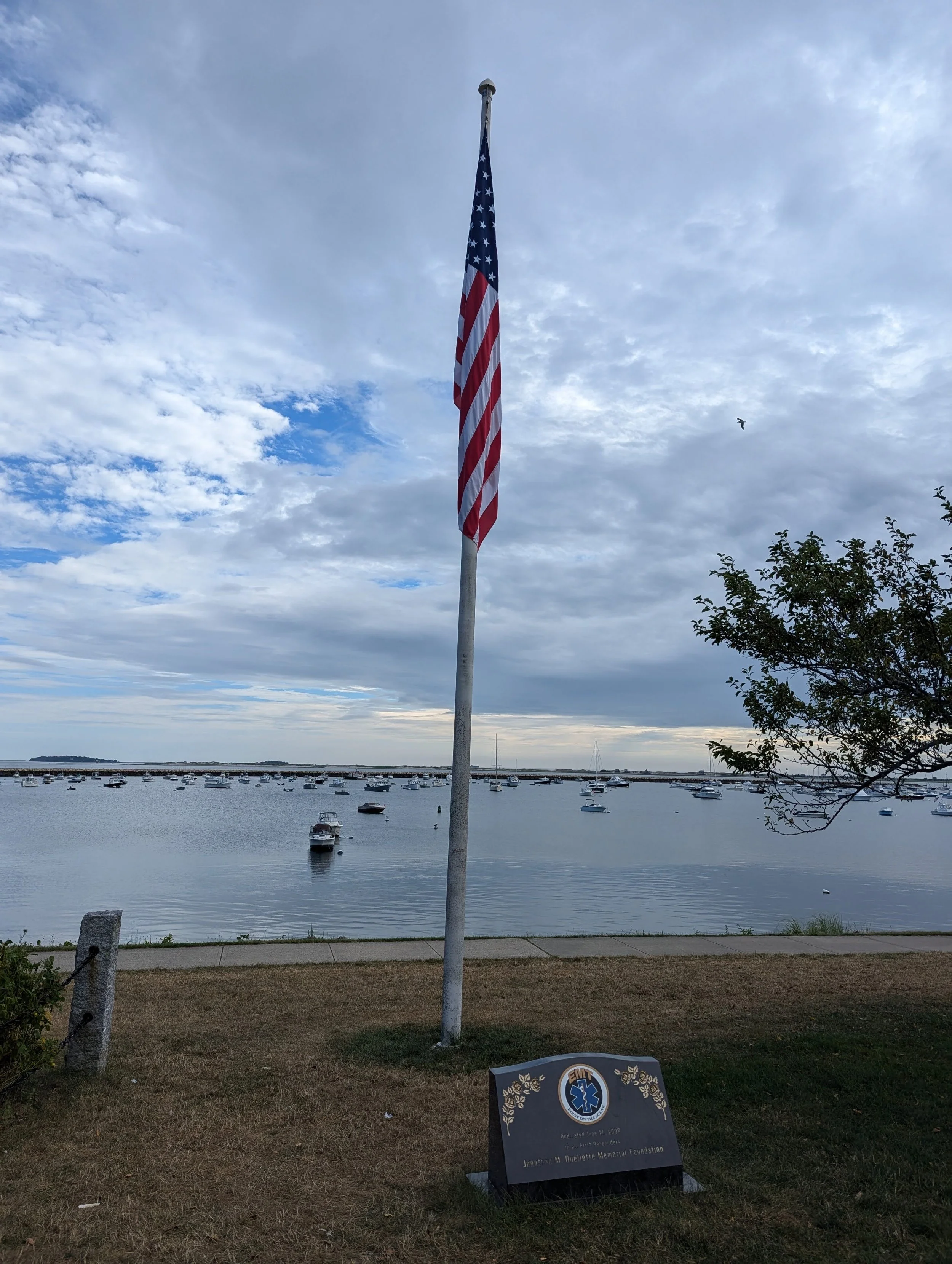 An American flag on a flagpole near a harbor with several boats anchored, a memorial plaque on the ground, a cloudy sky, and a tree on the right.