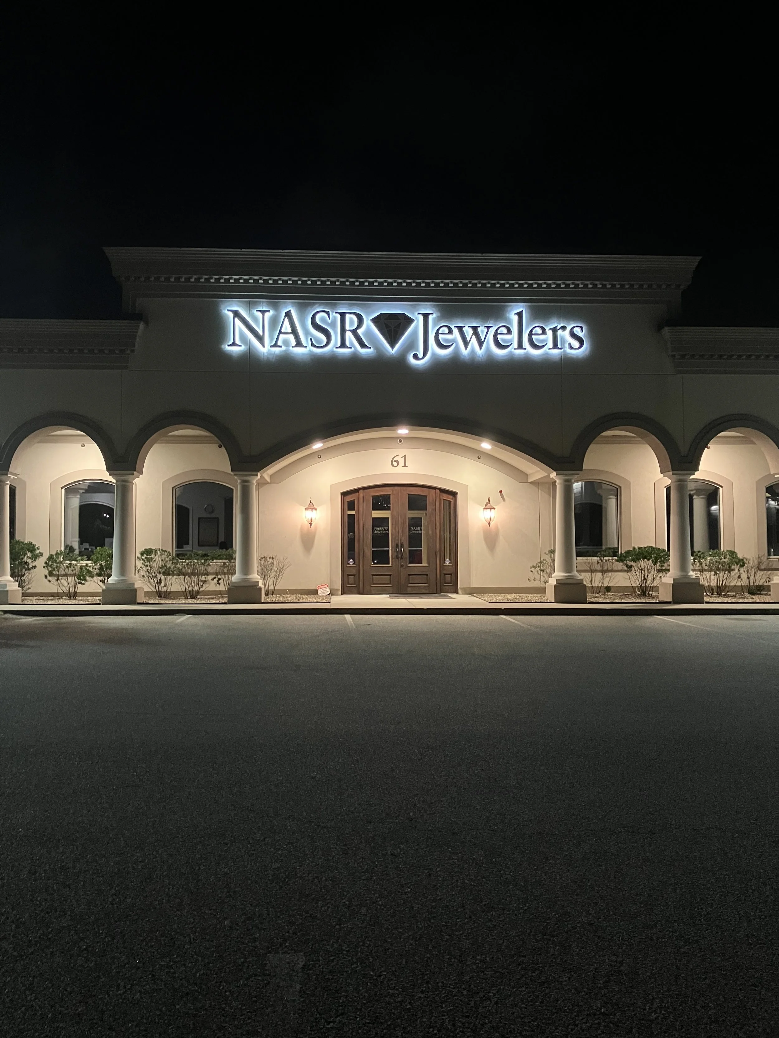 Night view of NASR Jewelers storefront with illuminated sign and front door, surrounded by landscaping