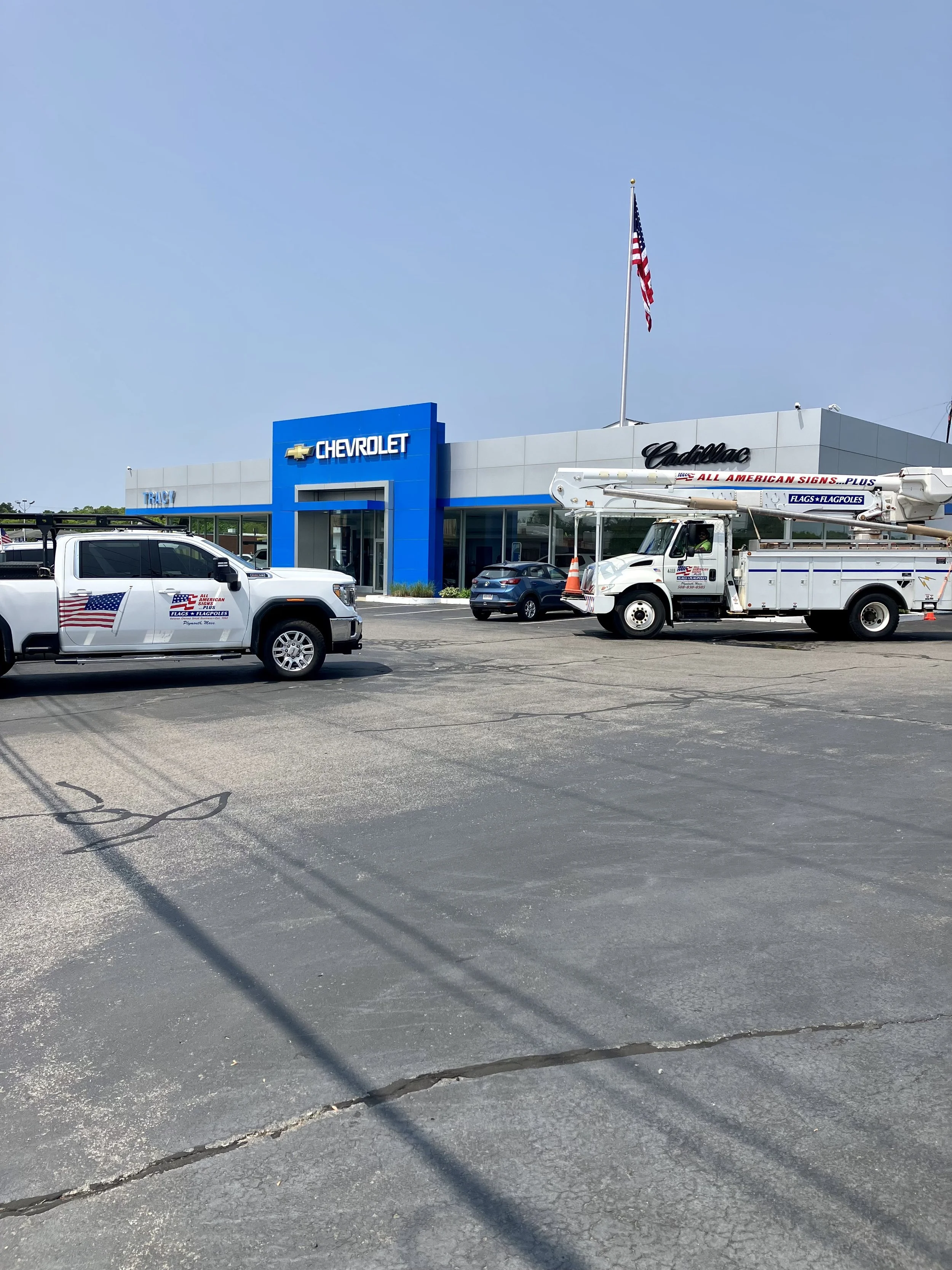 Car dealership with Chevrolet and Cadillac signs, American flag, service trucks, parking lot, blue sky.