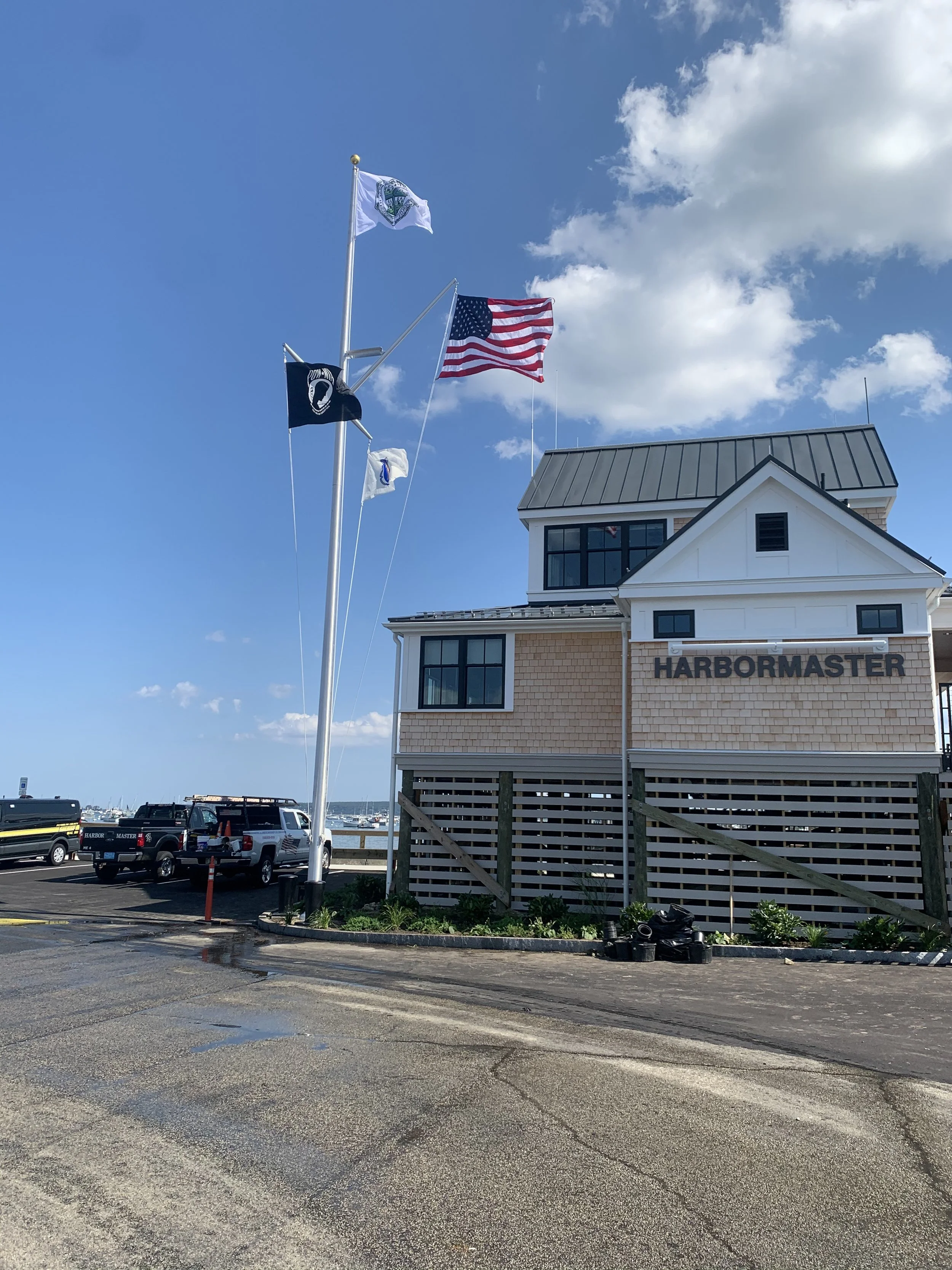 A building with a sign that reads 'HARBORMASTER' and flags flying on a tall flagpole, including the American flag, with a parking lot and boats visible in the background on a clear day.