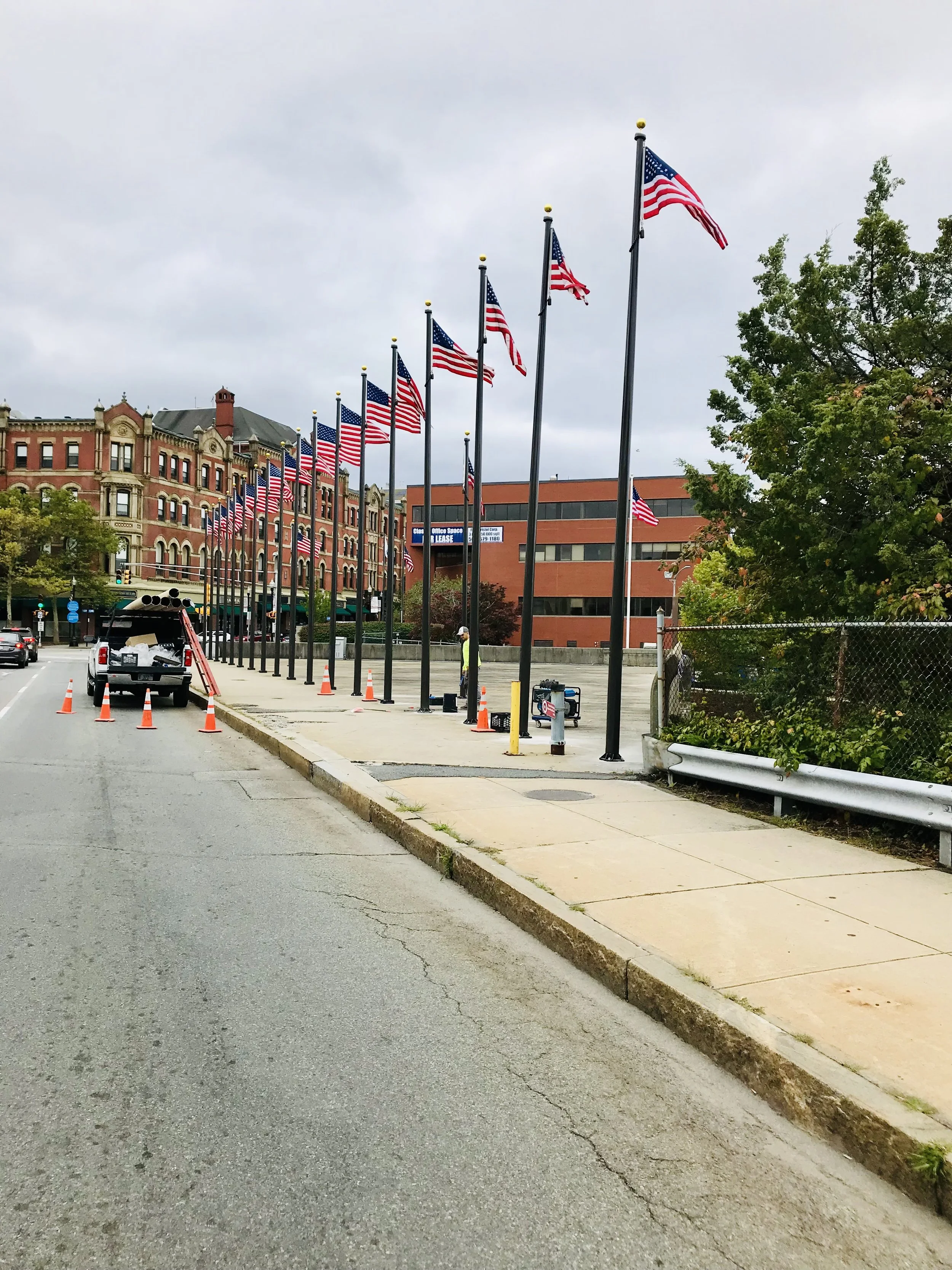 A row of American flags on tall poles along a sidewalk in an urban area with historic brick buildings, a construction area with cones, and a fence, under an overcast sky.