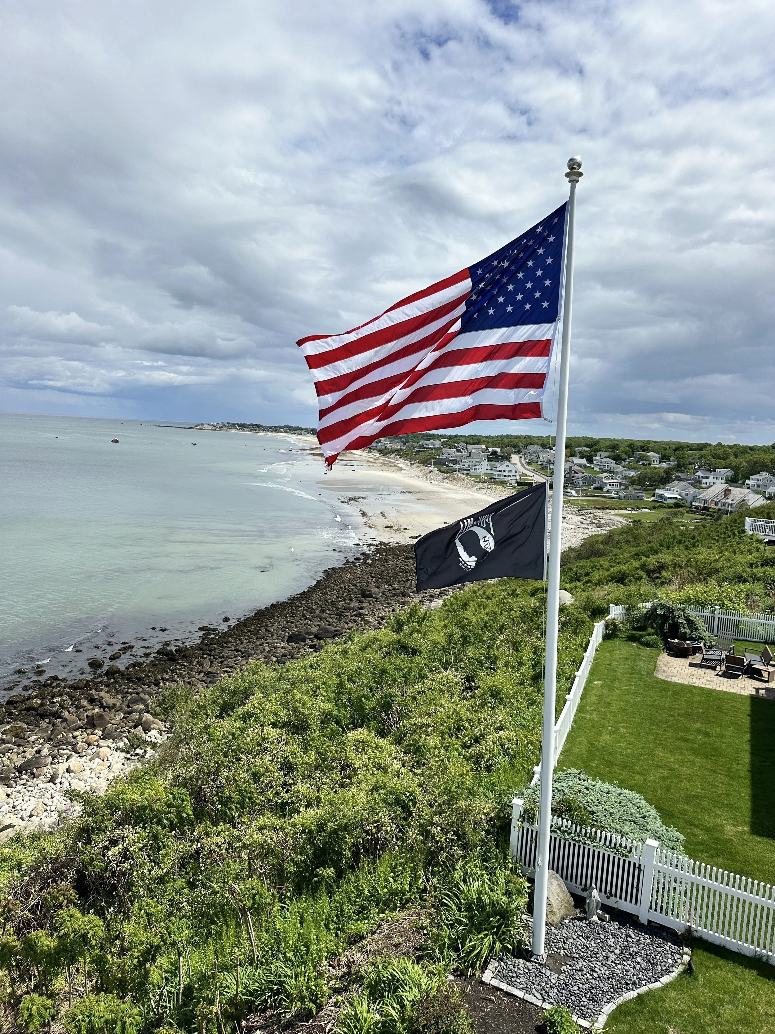American flag and Navy flag flying over a coastal residential area with a beach, ocean, and cloudy sky in the background.