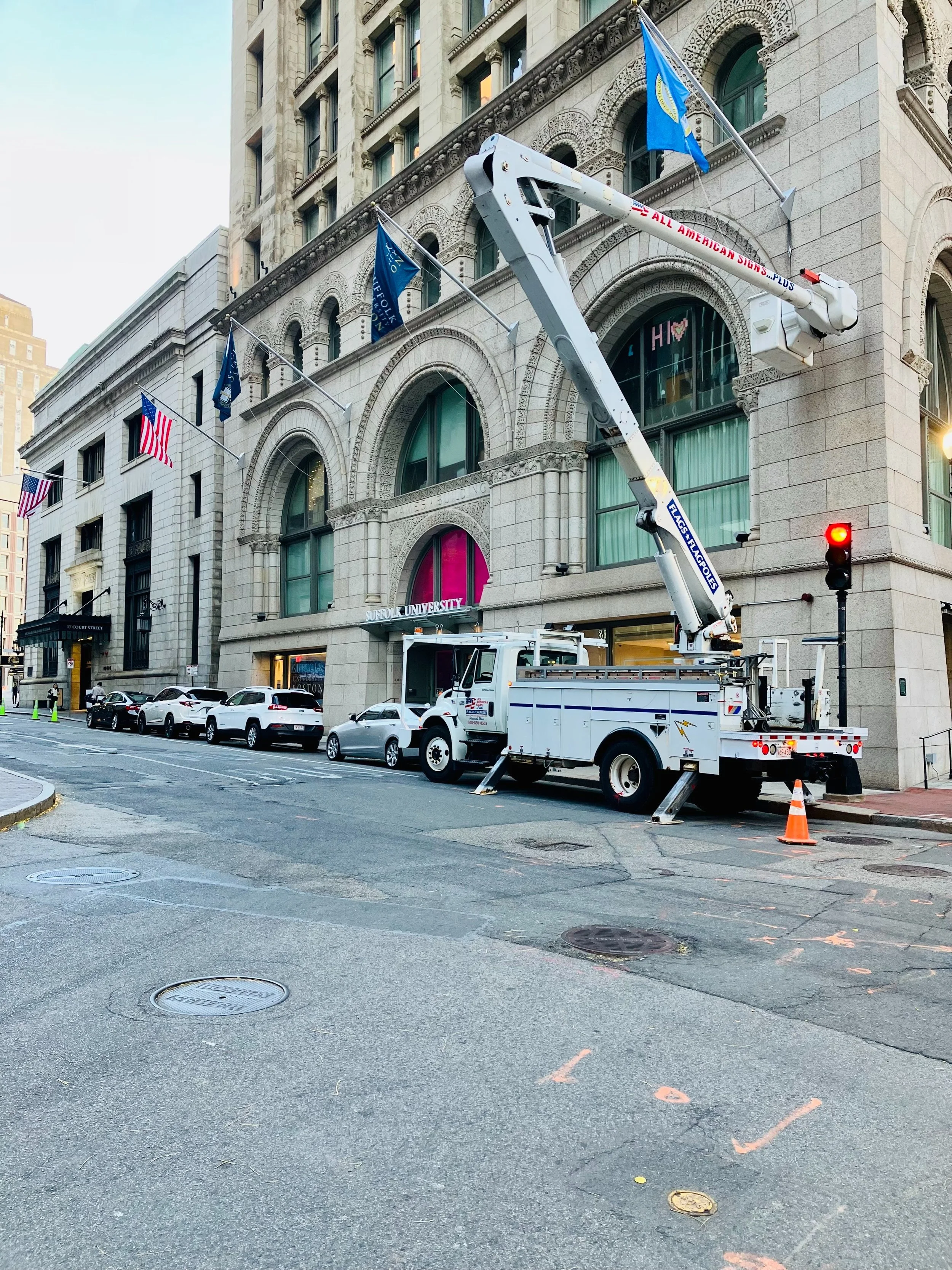 Bucket truck with a lift extended, working on the side of a historic building. Flags are hanging from the building, and there is a traffic light showing red. Several parked cars are along the street.