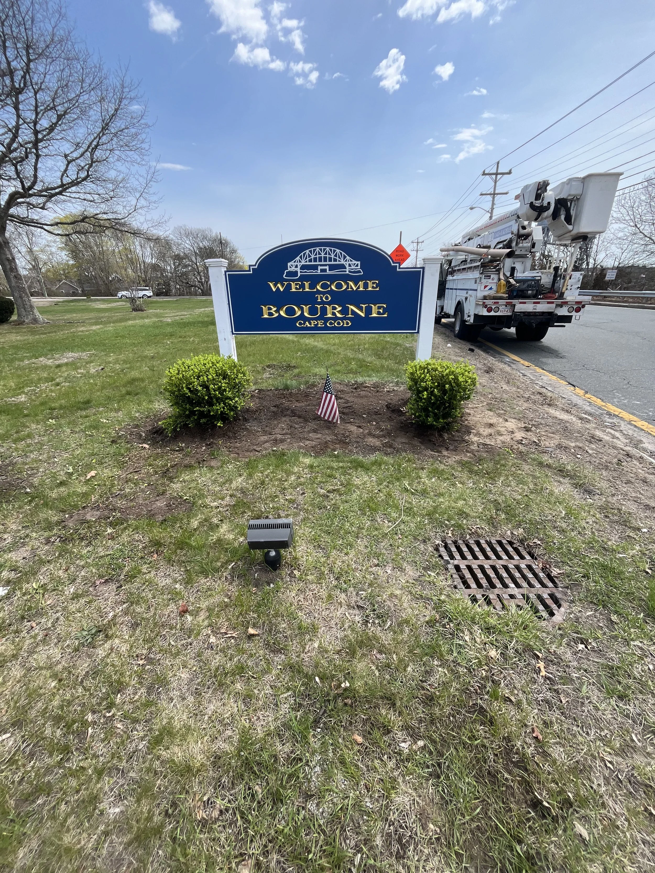 A welcome sign that reads 'Welcome to Bourne Cape Cod' in blue with gold lettering, flanked by two small green bushes, under a partly cloudy sky. There is a utility truck with a bucket lift parked on the street to the right, and a small American flag in front of the sign.