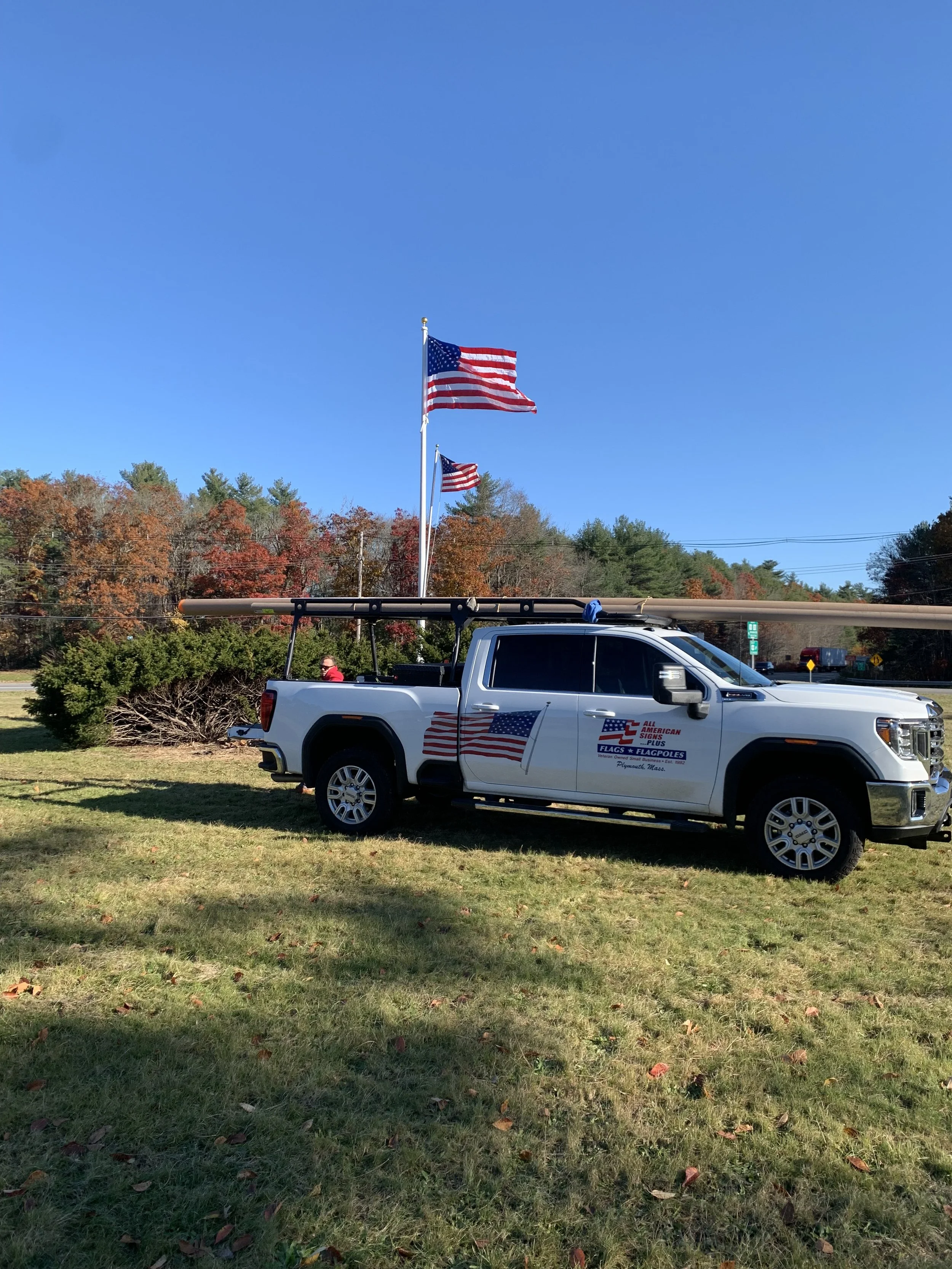 A white pickup truck with flags and a pole on its bed, parked on grass with American flags flying in the background and trees with autumn leaves.